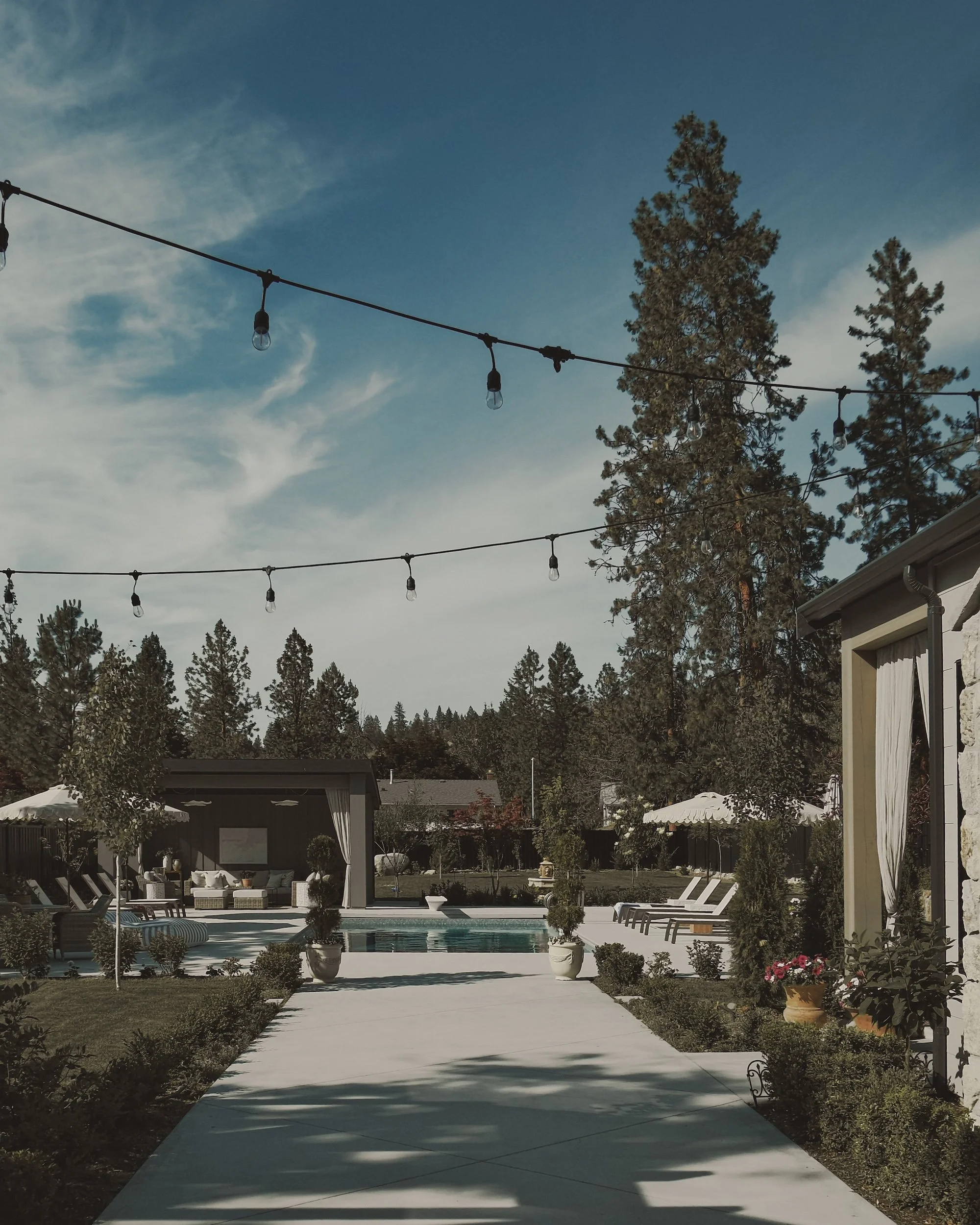 Outdoor backyard with a swimming pool, surrounded by lounge chairs, potted plants, and trees, under a partly cloudy sky with string lights overhead.