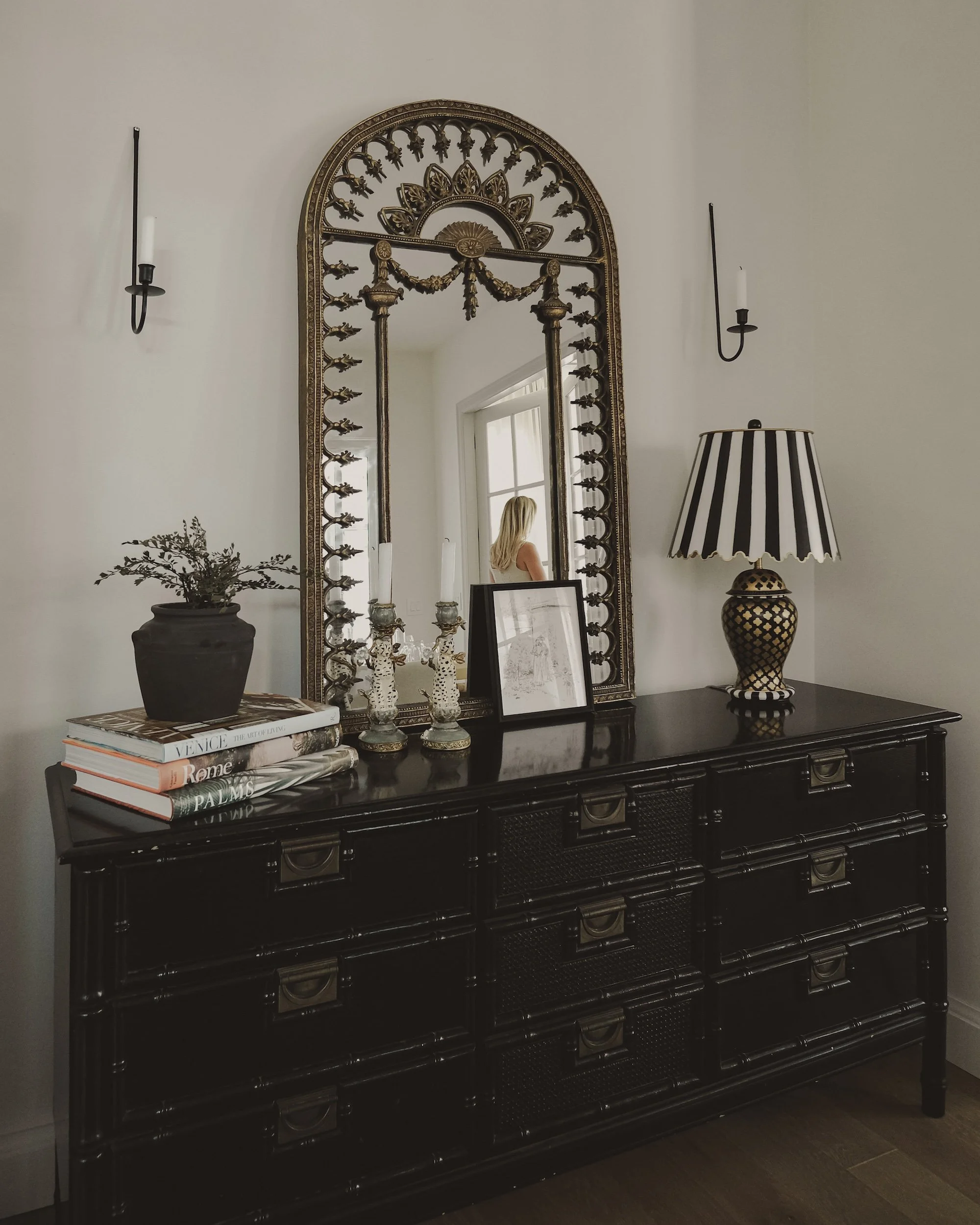 Dark wooden dresser with a large ornate mirror above, decorated with books, small framed photos, a plant, a table lamp, and candlesticks.