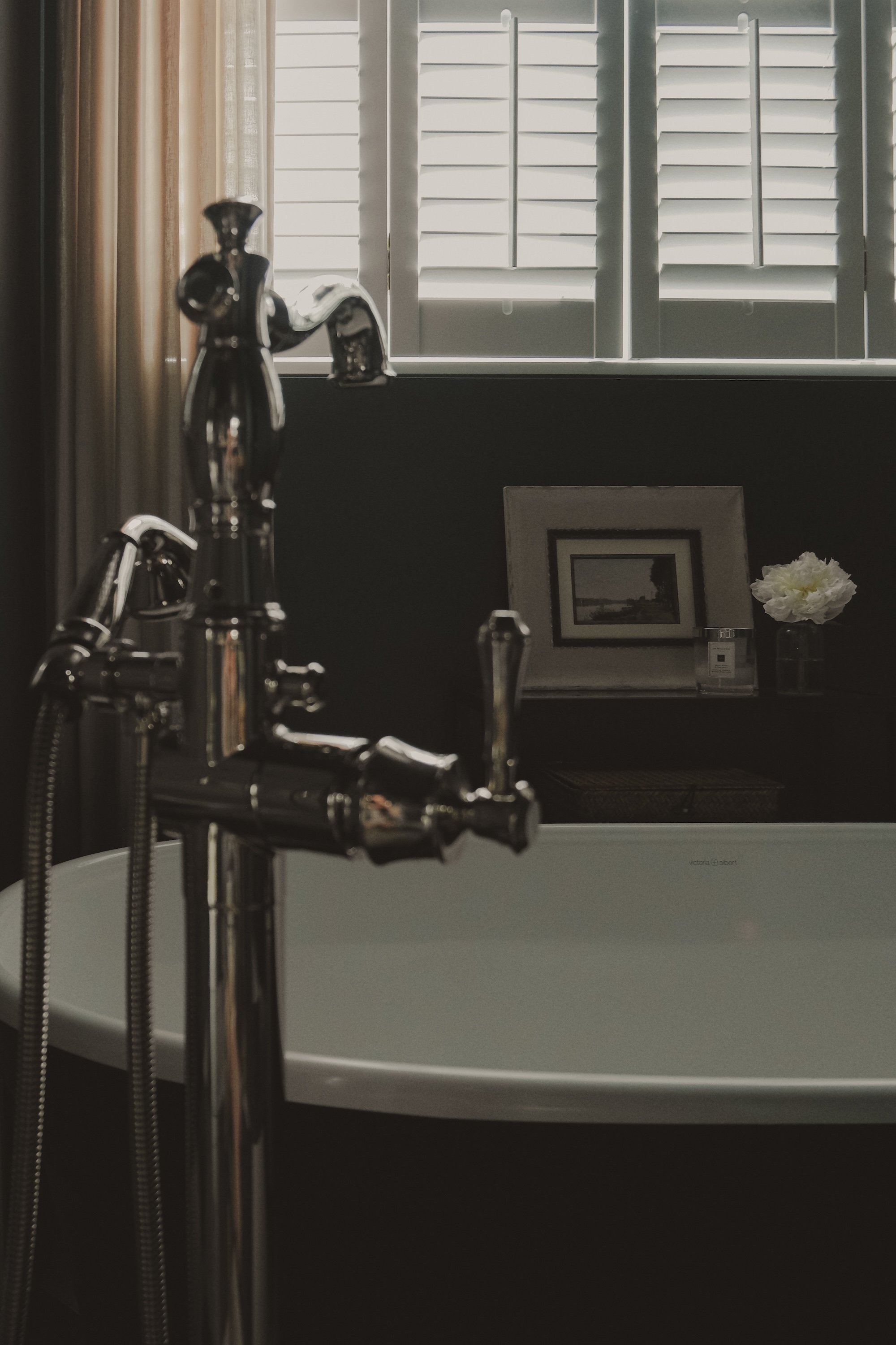 Bathroom with a white bathtub, silver faucet, window with shutters, and a small shelf with framed photo, flower, and candle.