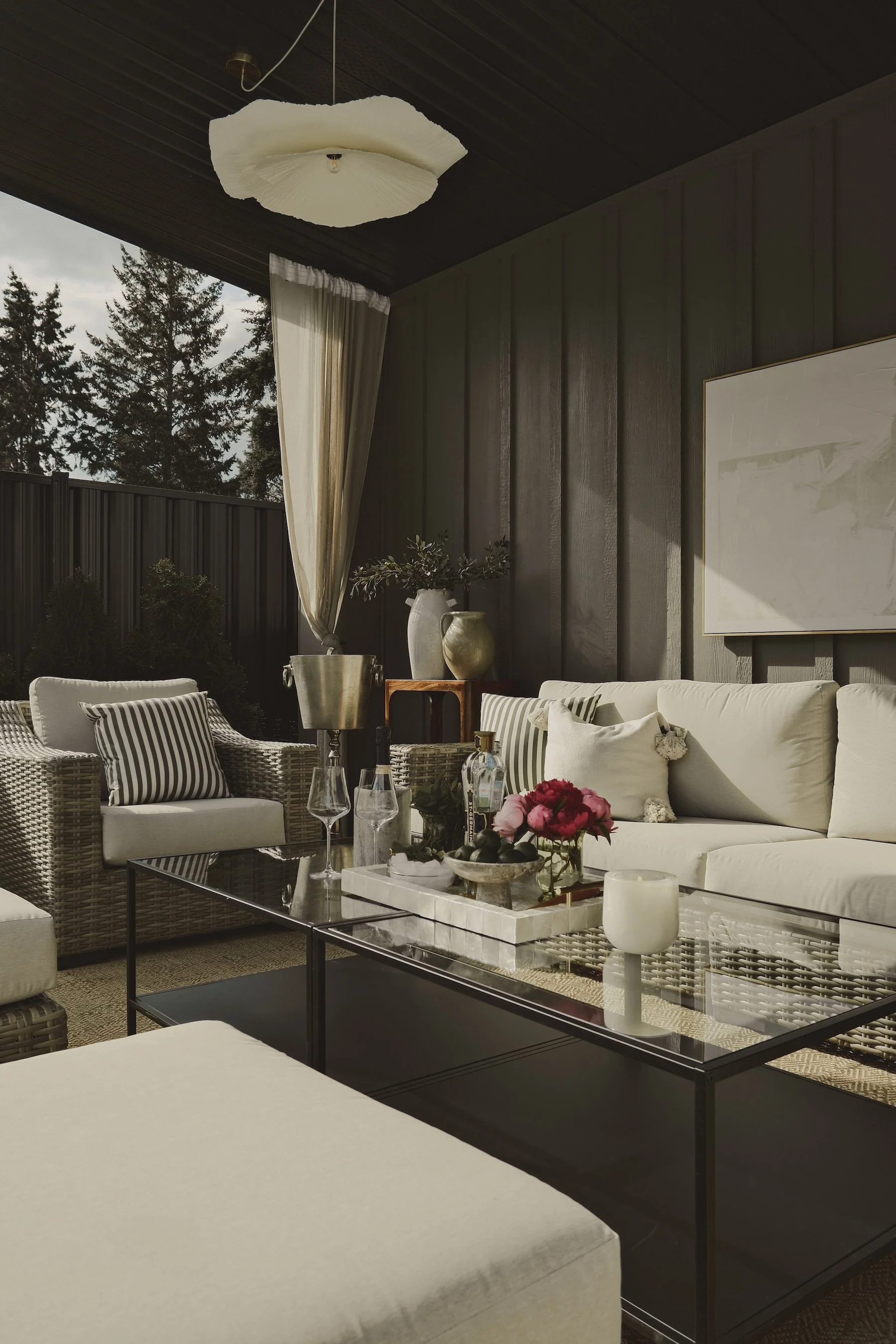 Outdoor patio with wicker sofas, a glass coffee table, and decorative pillows. The table has flowers, glassware, and a tray. The space features dark wood paneling, curtains, and potted plants, with trees visible outside.