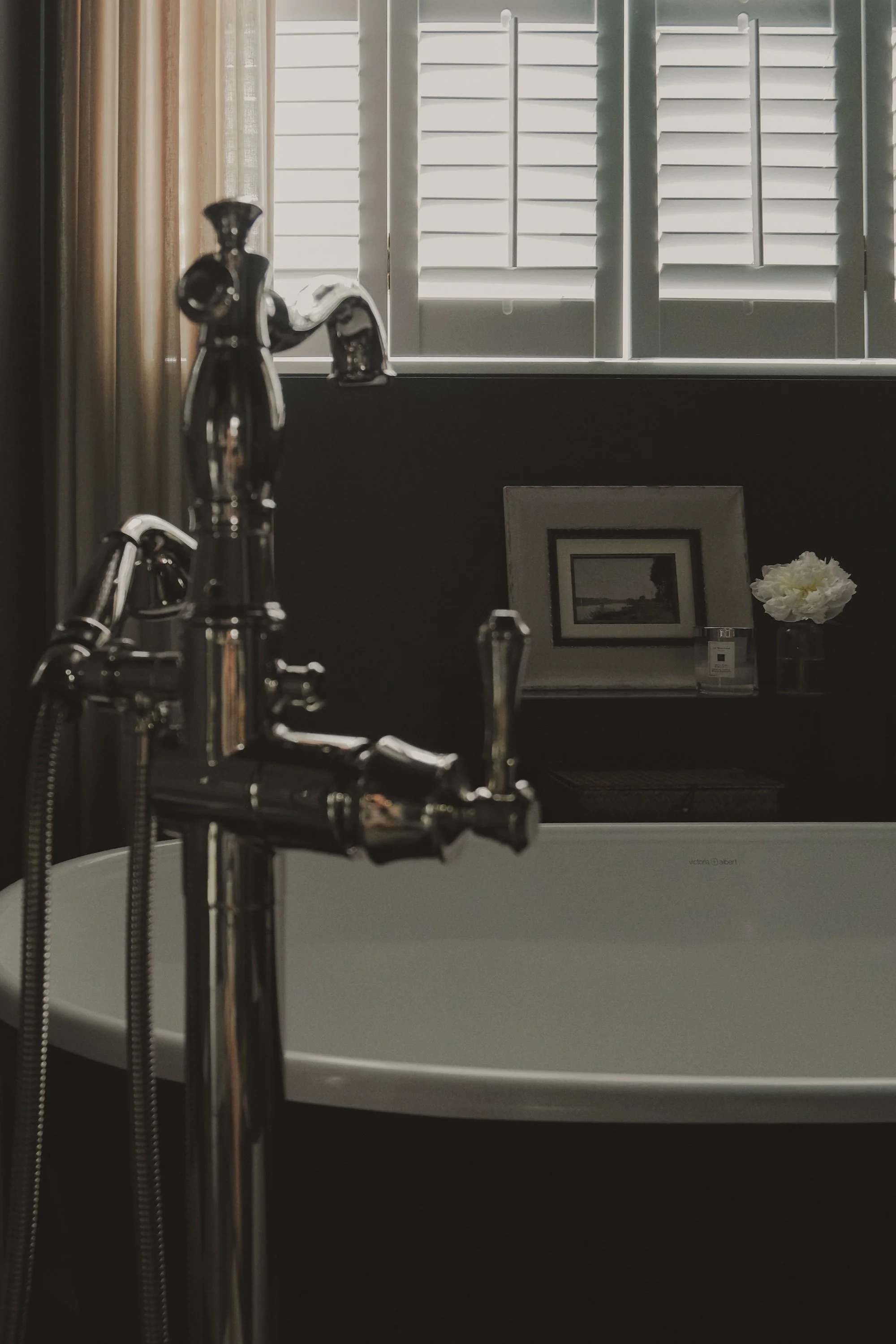 A bathroom with a chrome faucet and a white bathtub, a framed picture, a candle, and a flower in a vase on a shelf, with light coming through plantation shutters.