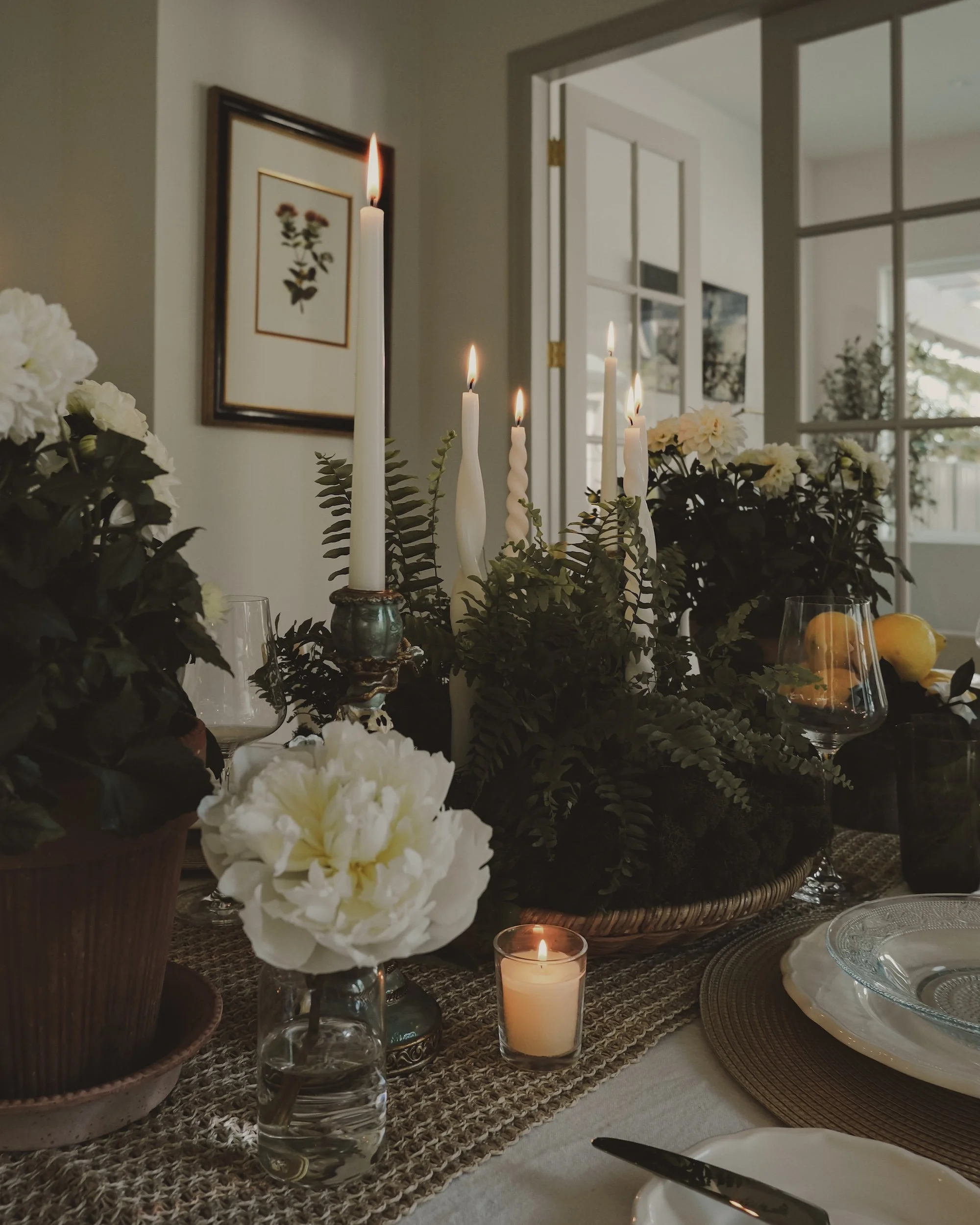 A dining table decorated with candles, white flowers, and greenery in a well-lit room, with a window and artwork on the wall in the background.