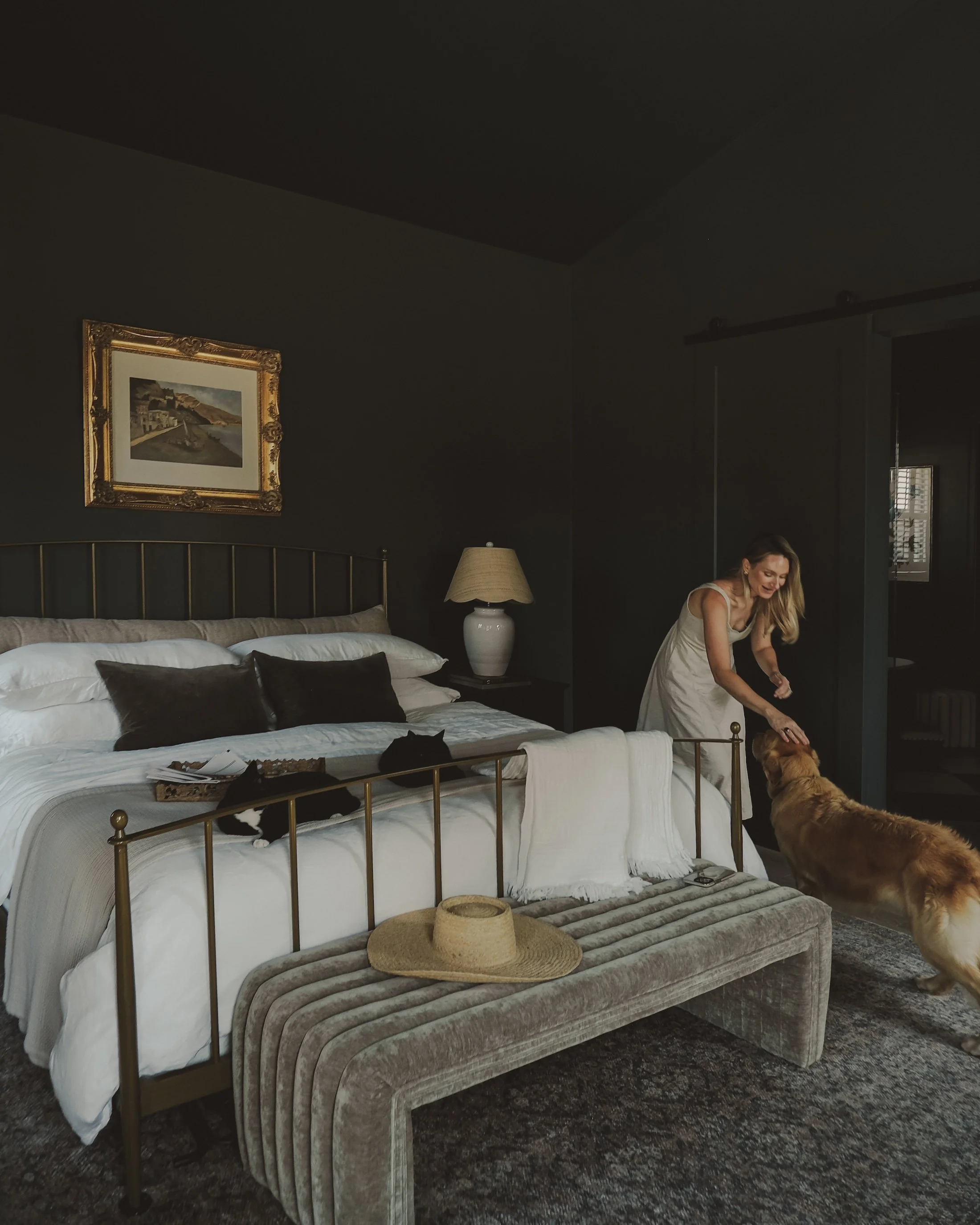 Woman playing with a dog in a dark-colored bedroom with a bed, pillows, and a bench at the foot of the bed.