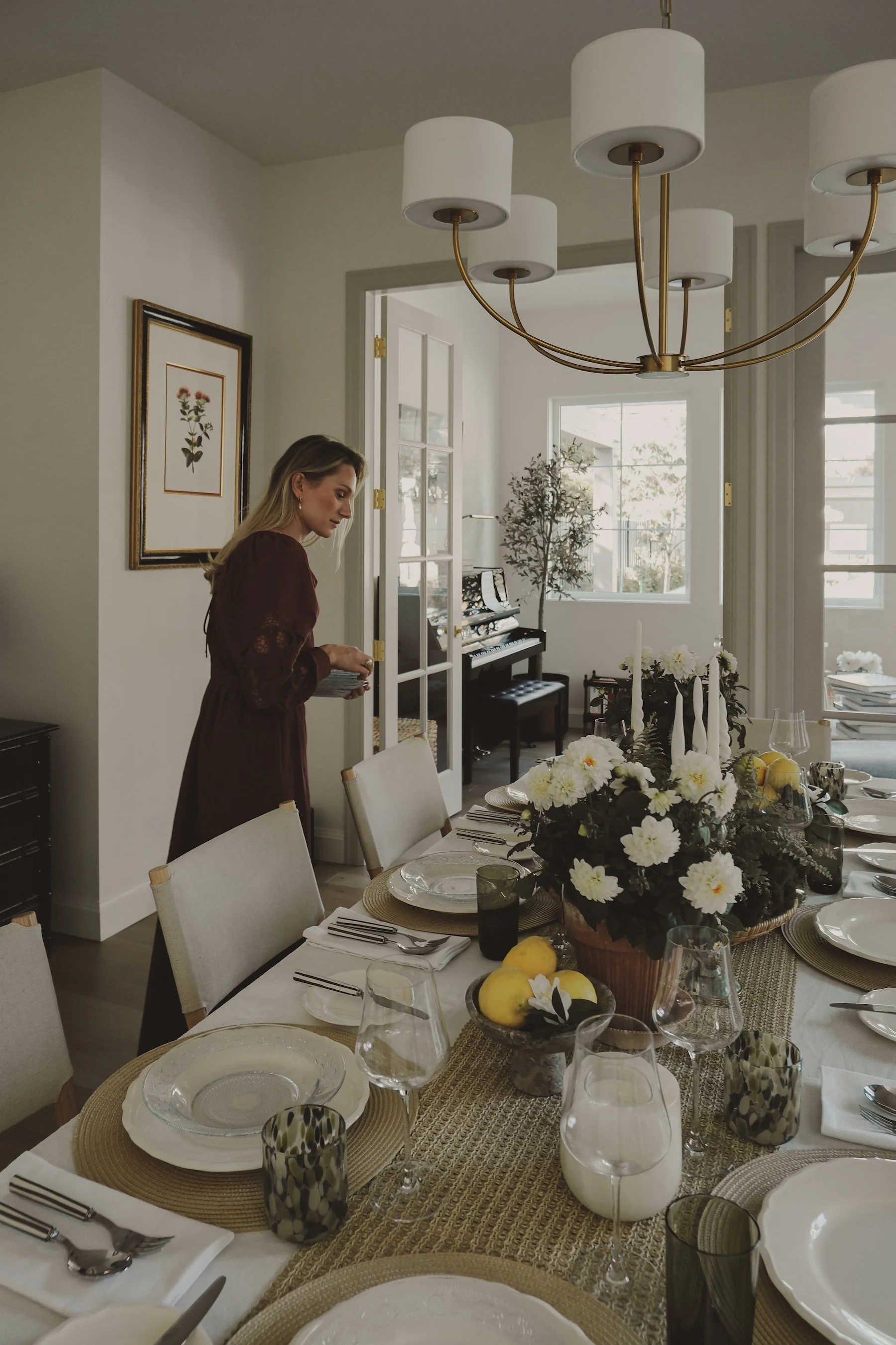 A woman in a maroon dress stands near a dining table set with plates, glasses, and napkins, in a bright room with a chandelier and framed artwork on the wall.