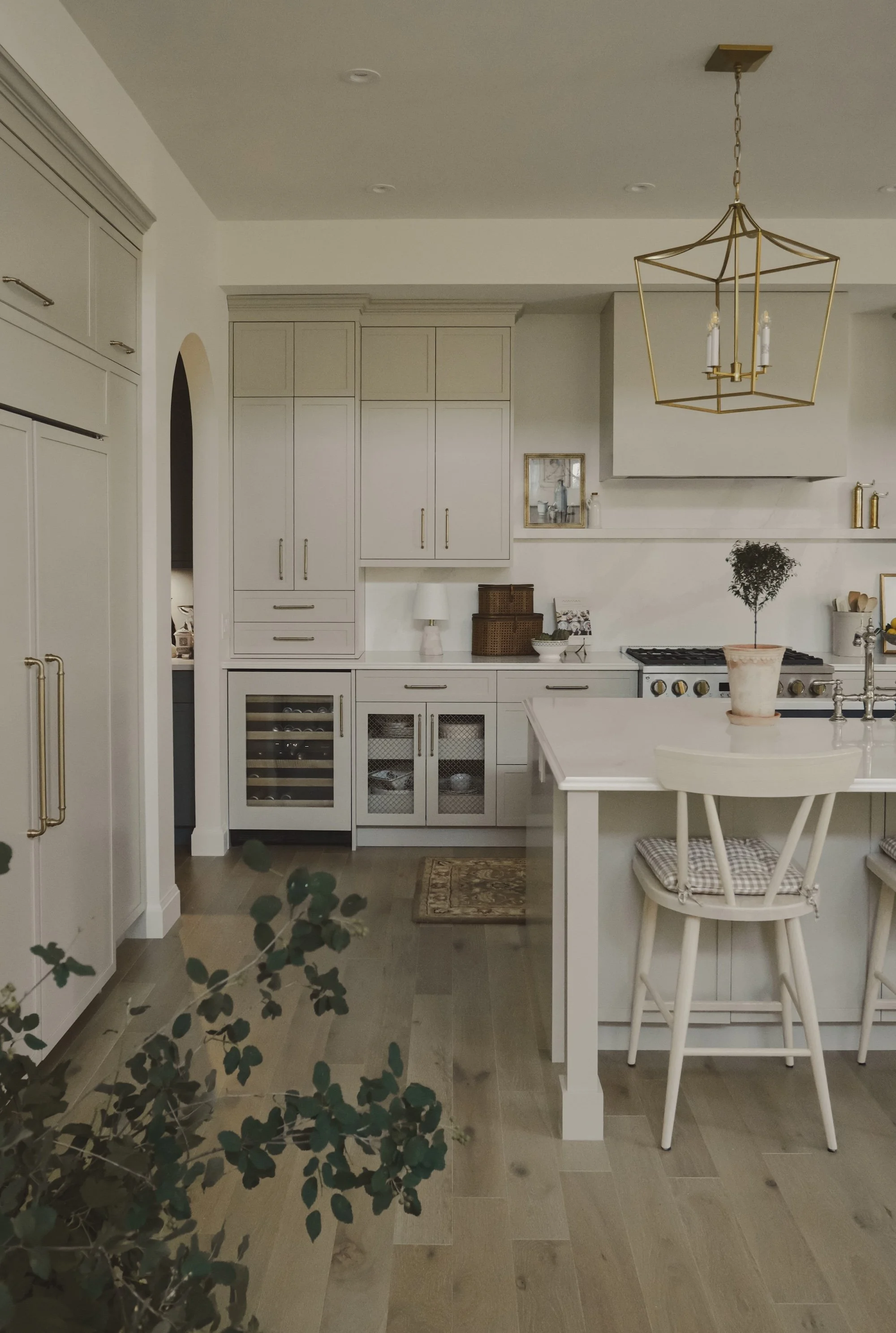 Modern white kitchen with light wood flooring, gold accents, and minimalist decor, including a white island, cabinets, and a hanging gold geometric chandelier.