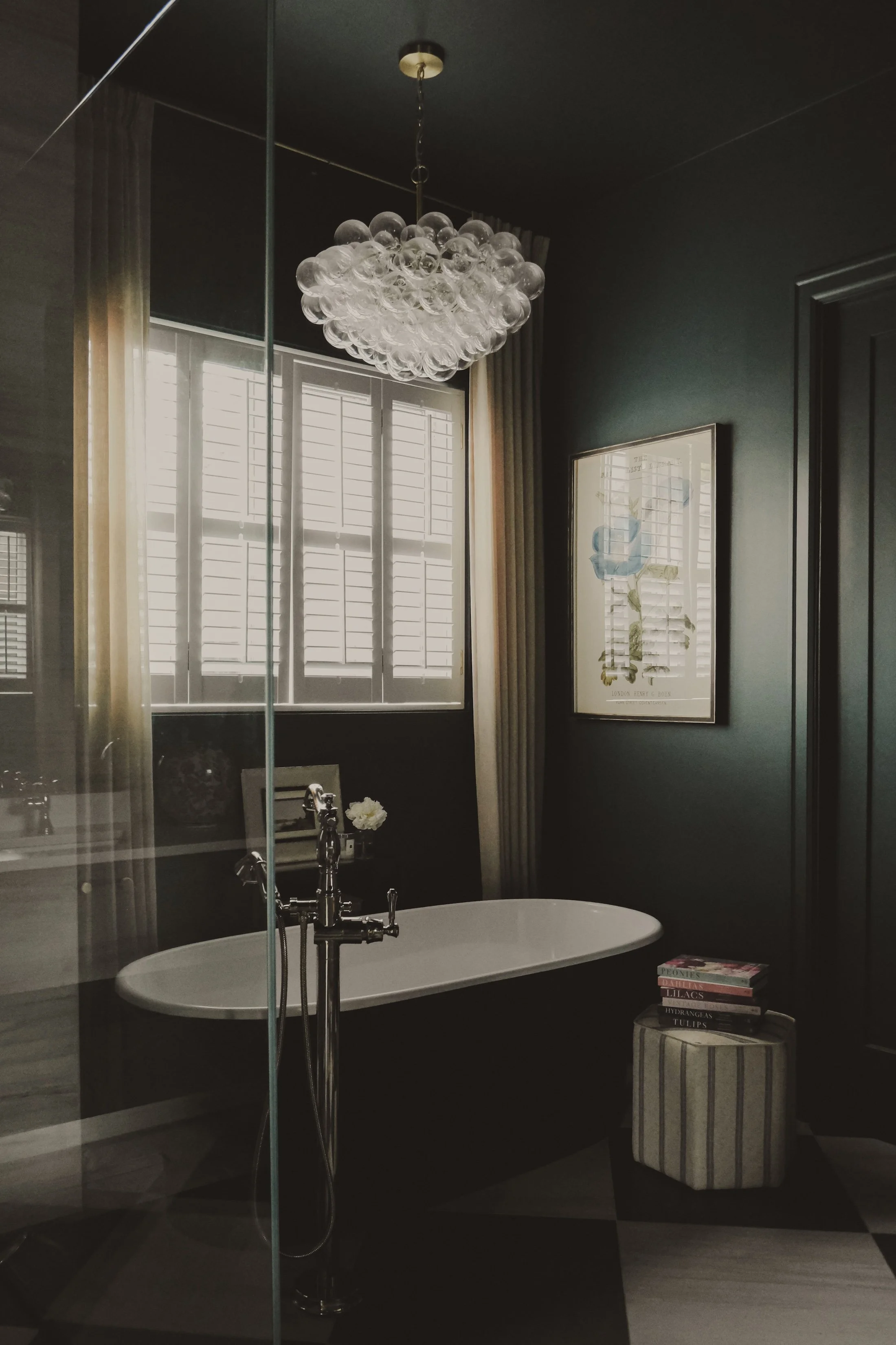Elegant bathroom with a black bathtub, large window with shutters, framed artwork, striped ottoman with stacked books, and a modern glass chandelier.