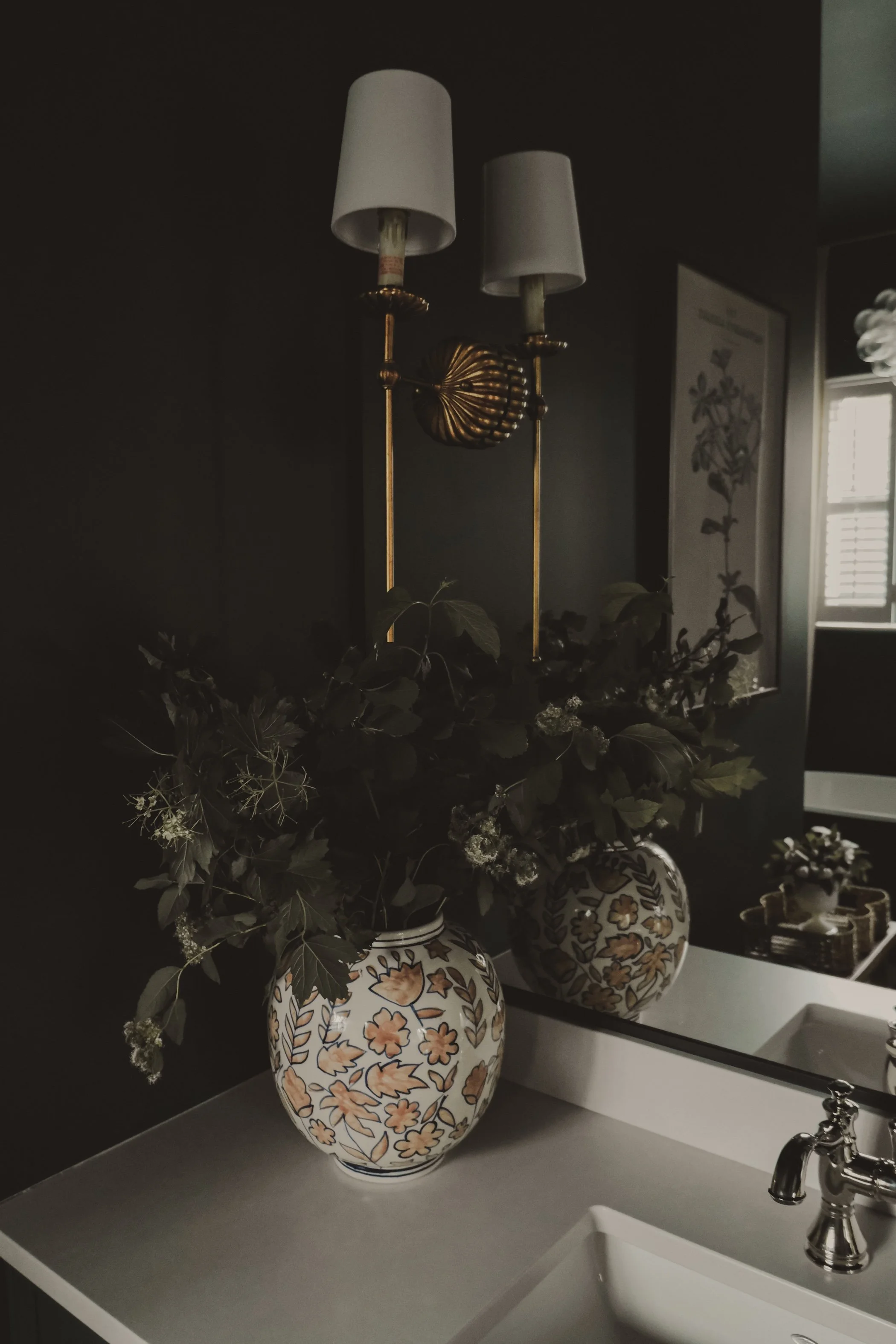 A bathroom counter with a ceramic vase holding greenery, a mirror, a sink, and a wall-mounted light fixture with two lamps.