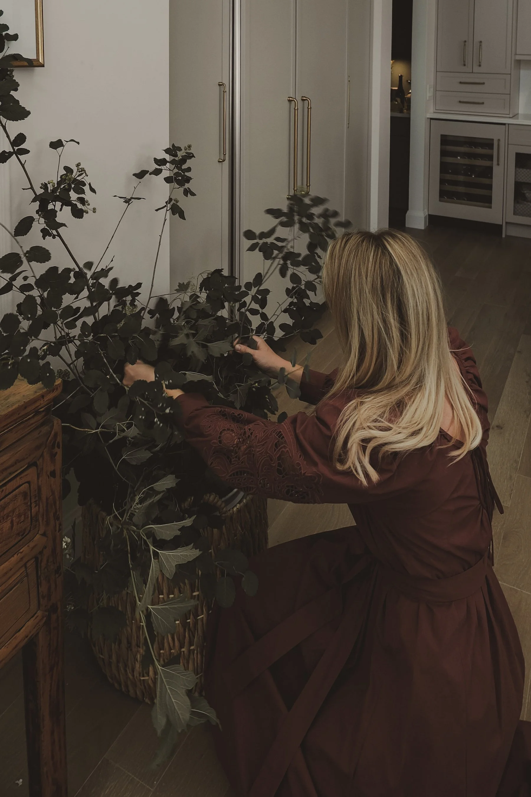 A woman with blonde hair wearing a burgundy dress arranging or handling a large dark green leafy plant inside a basket in a modern kitchen.