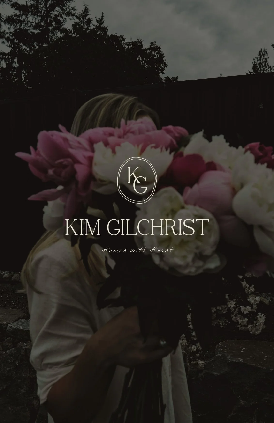 A woman holding a large bouquet of pink and white flowers in front of her face, with a dark outdoor background and the text 'Kim Gilchrist Homes with Heart' in the foreground.