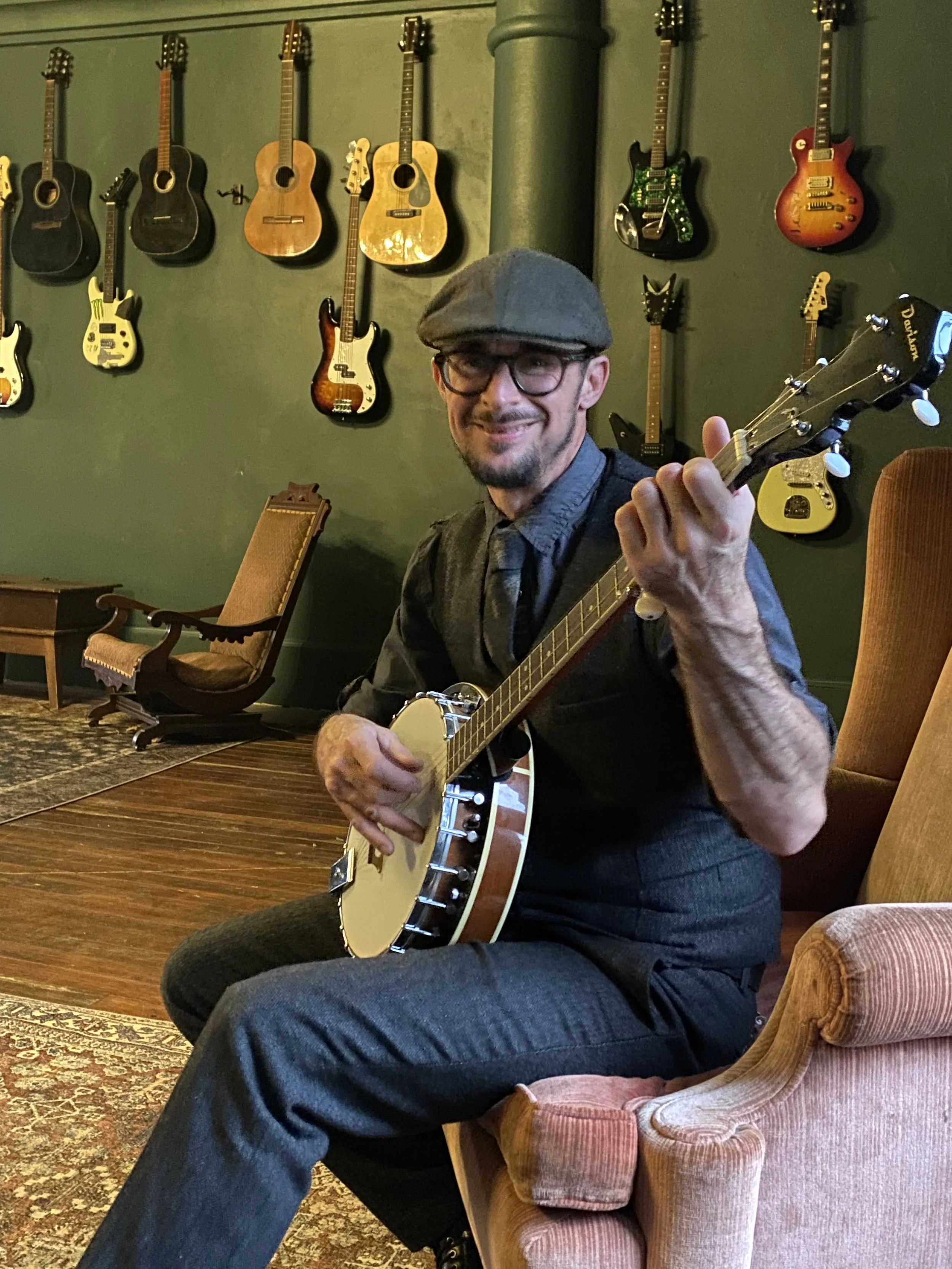 A man in a gray hat and glasses playing a banjo in a room with green walls and a collection of guitars hanging on the wall behind him.