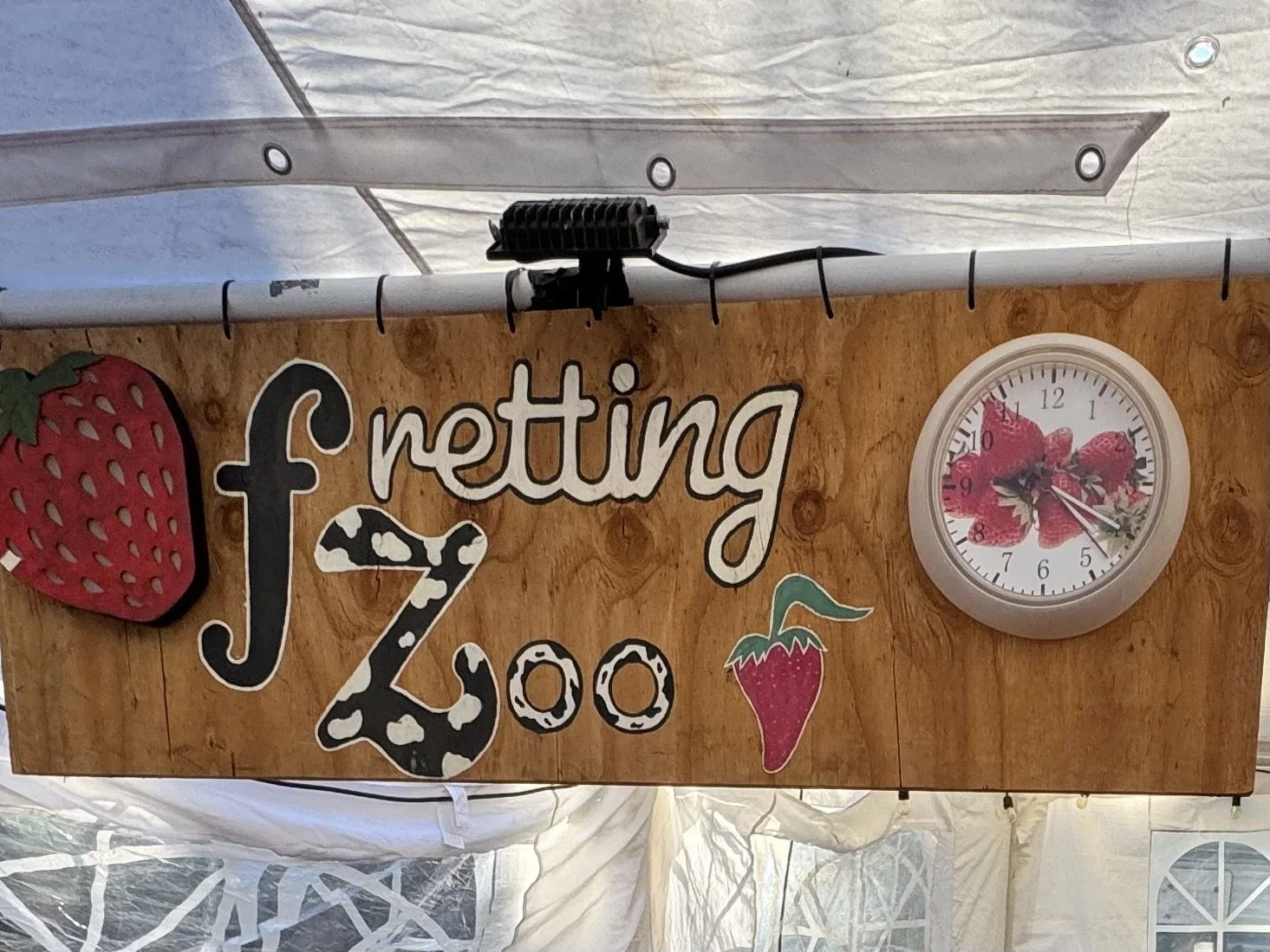 A wooden sign with the words "Fretting Zoe" and strawberry-themed decorations, including a strawberry illustration and a strawberry-shaped clock showing around 5:24. Part of a strawberry decoration is visible on the left.
