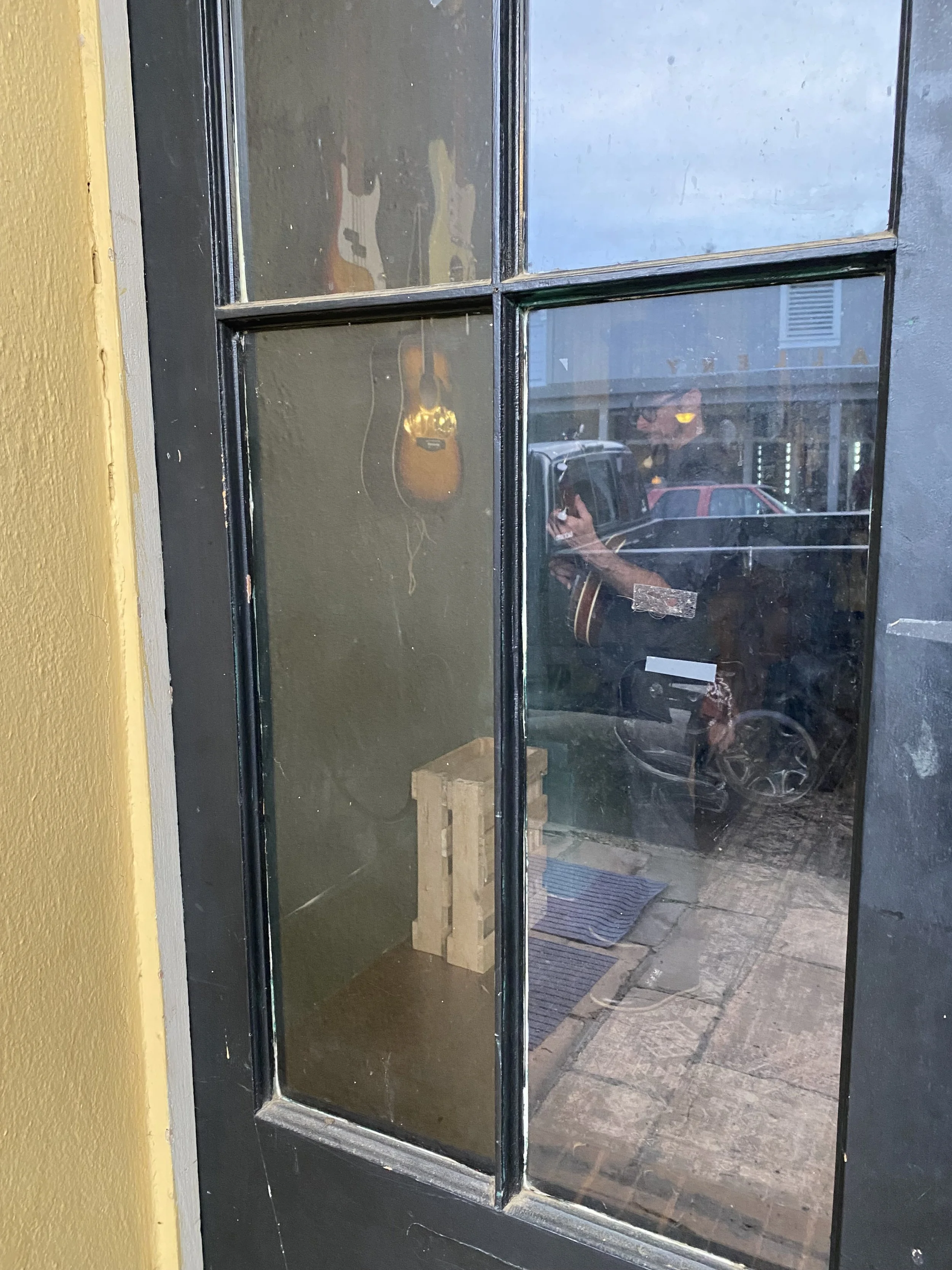 Looking through a glass window with four panes, showing reflections and a partial view of a person taking the photo, a guitar hanging on the wall, a wooden pallet on the floor, and parked cars outside.