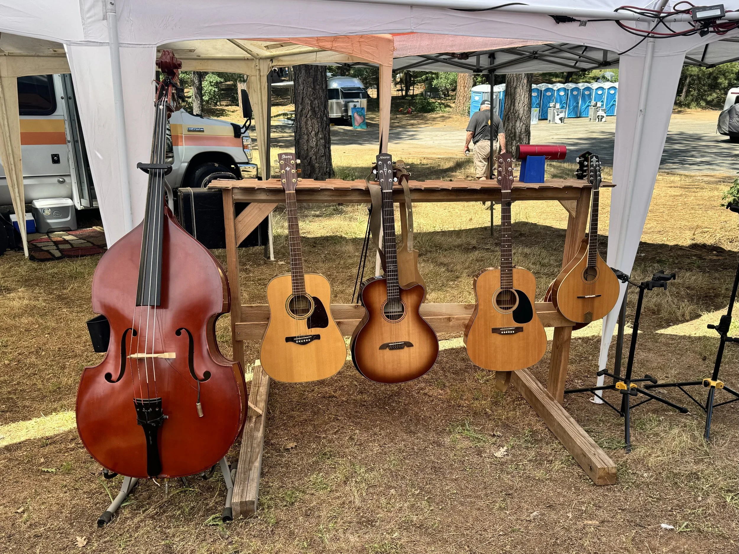 A display of five stringed musical instruments, including a large upright bass and four acoustic guitars, hanging on a wooden stand under a white outdoor tent at an outdoor event.