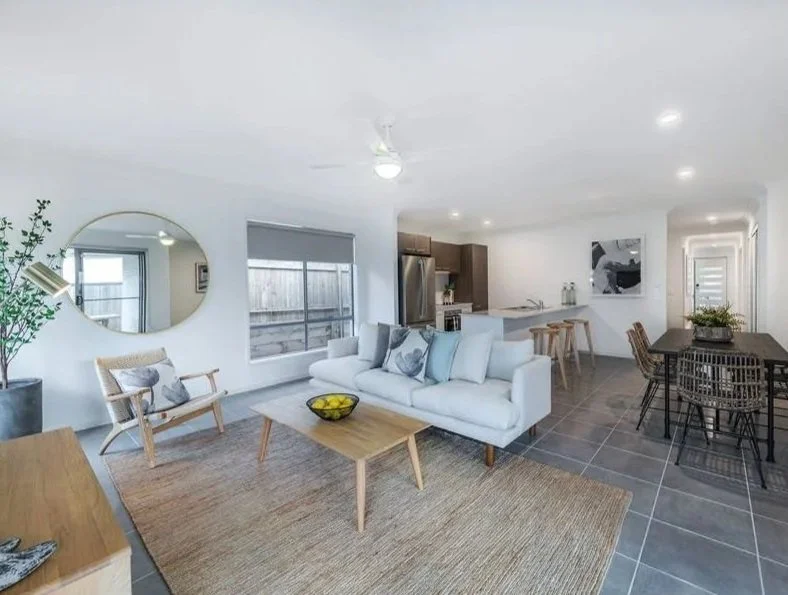 Modern open-concept living room and kitchen with white walls, large windows, light-colored furniture, a wooden coffee table, black dining table with chairs, and gray tiled flooring.