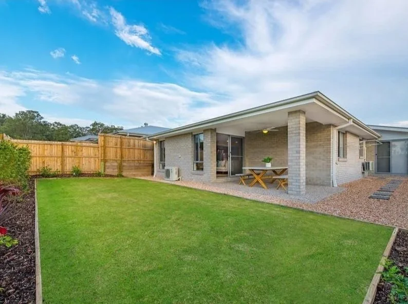 Backyard of a modern house with a covered patio, green lawn, and wooden fence under a blue sky with clouds.