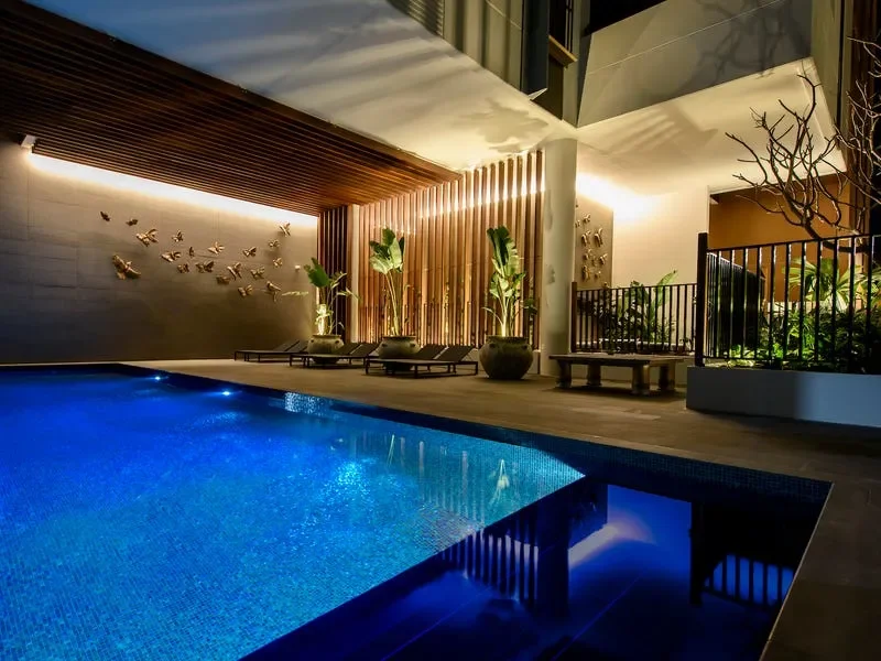 Modern outdoor swimming pool area at night with wooden decor, potted plants, and outdoor seating.