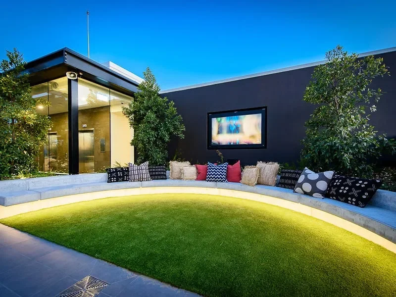 Outdoor lounge area with a curved concrete bench, colorful pillows, green grass, two trees, a large black wall with a mounted TV, and a modern building with glass walls and an elevator to the left