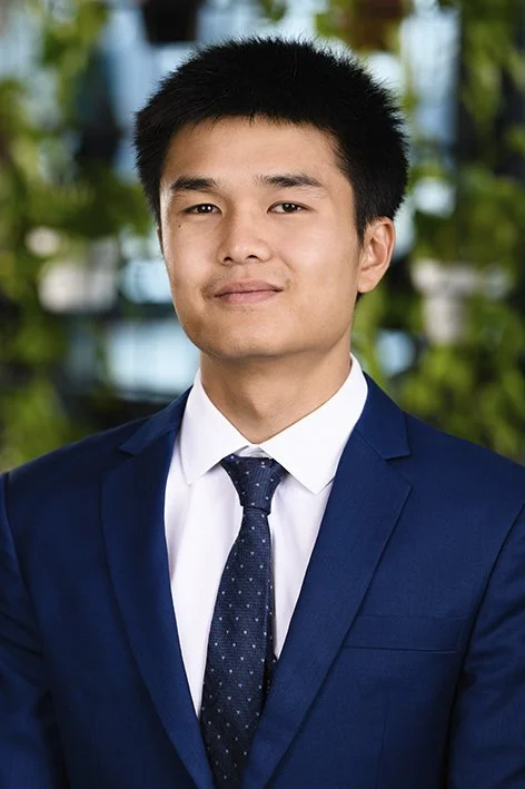 A young man in a navy suit, white shirt, and polka dot tie, standing outdoors with blurred green foliage in the background.
