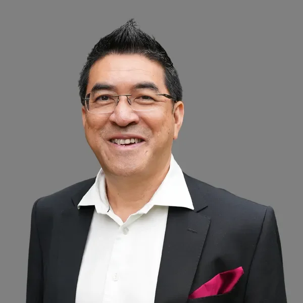A man with glasses smiling, dressed in a black suit with a white shirt and pink pocket square, against a plain gray background.