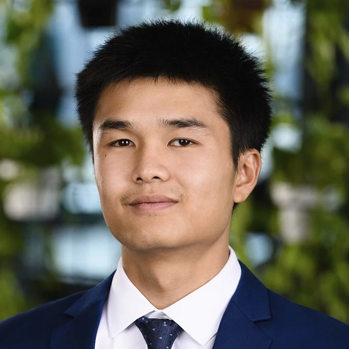 Professional portrait of a young man with short black hair, wearing a navy suit, white shirt, and dark tie, outdoors with blurred greenery in the background.