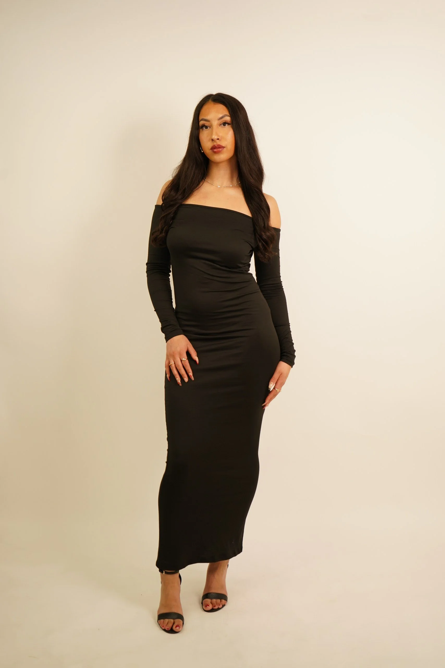 A woman with long dark hair in a black off-shoulder dress and heels standing against a plain light-colored background.