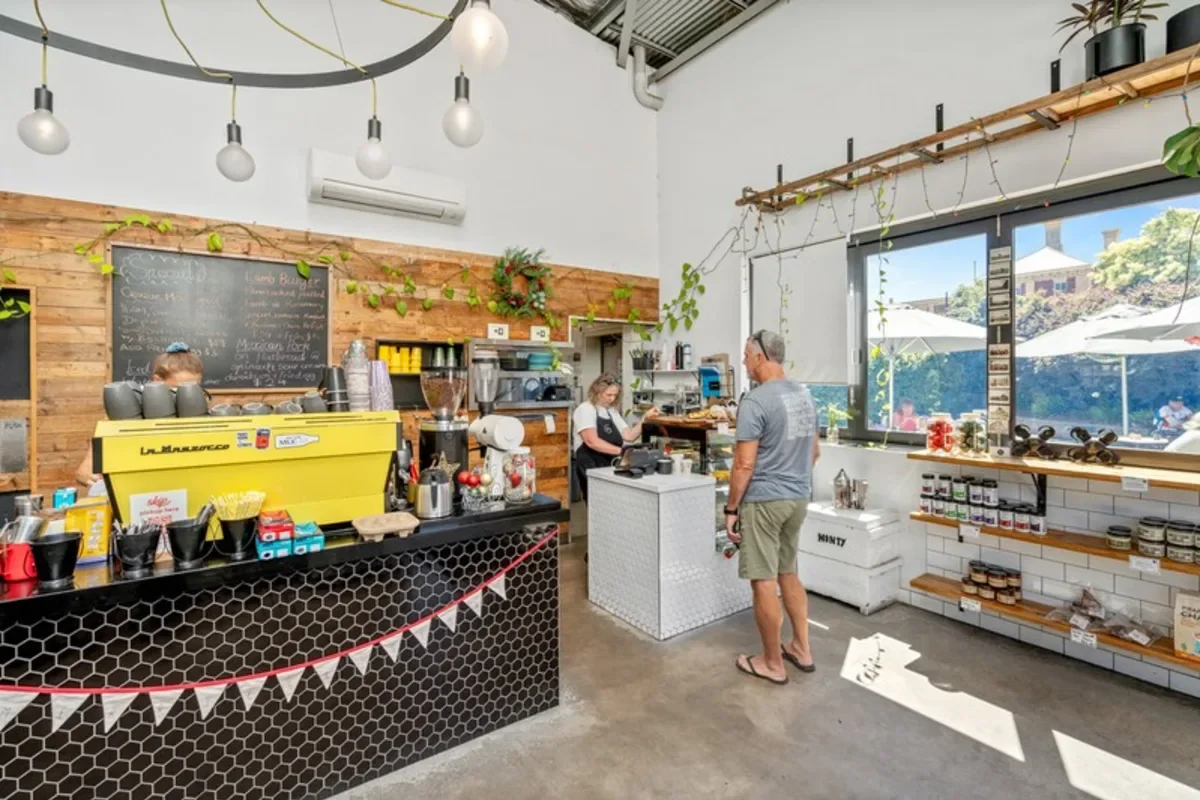 Inside a modern coffee shop with customers ordering at the counter, wooden accents, string lights, and large windows letting in natural light, with shelves displaying products