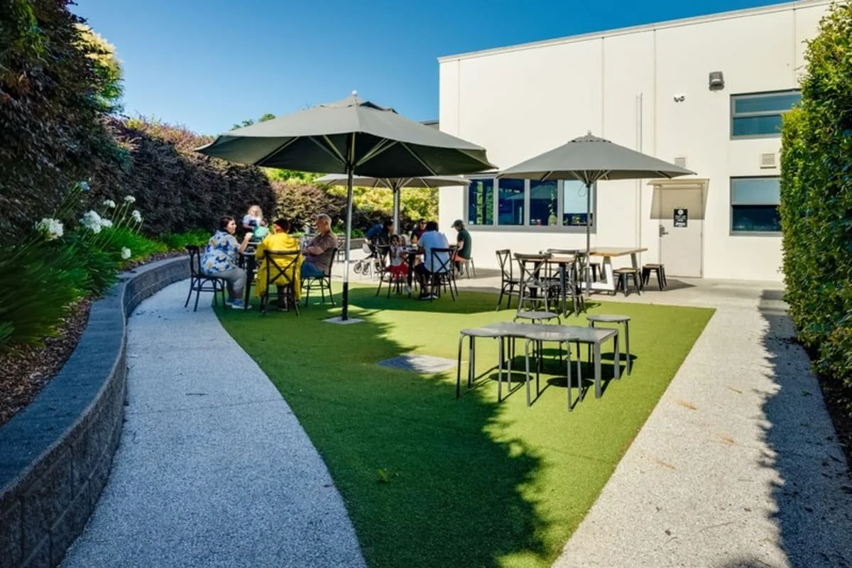 Outdoor patio area with tables, chairs, and umbrellas outside a modern building, with people sitting and socializing on a sunny day.