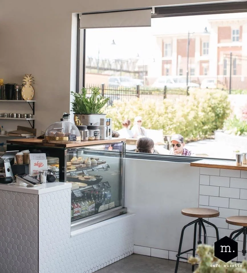 Interior of a cafe with a glass display case filled with pastries and drinks, a counter with a cash register, and a large window showing outdoor seating with people, plants, and buildings.