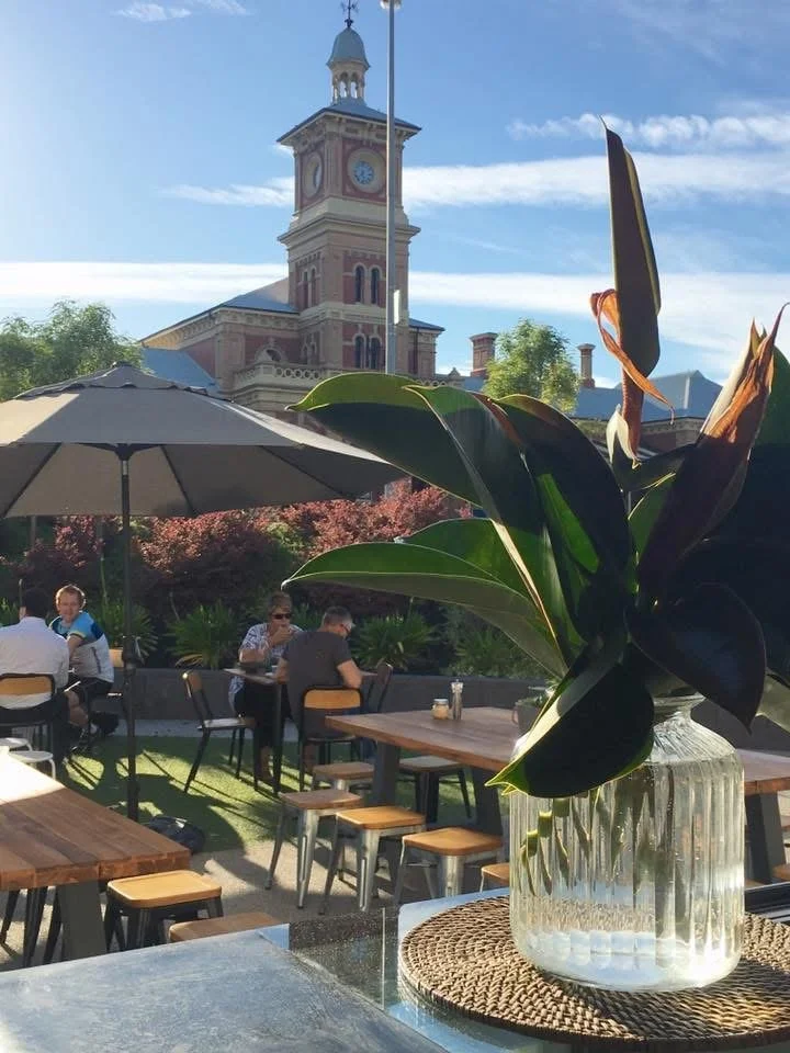 View of an outdoor cafe with people sitting at tables under large umbrellas, in front of a historic clock tower with a blue sky and some trees around.