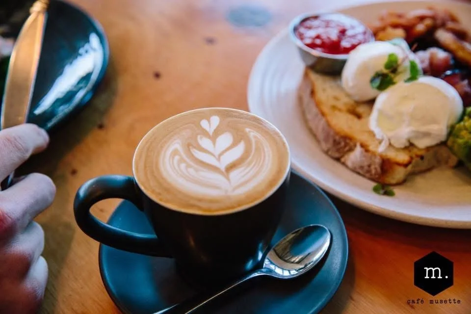 A black coffee cup with latte art on top, placed on a matching saucer with a small spoon. In the background, there's a plate with toast, topped with butter and a dollop of sour cream, accompanied by a small cup of red jam, and some vegetables.