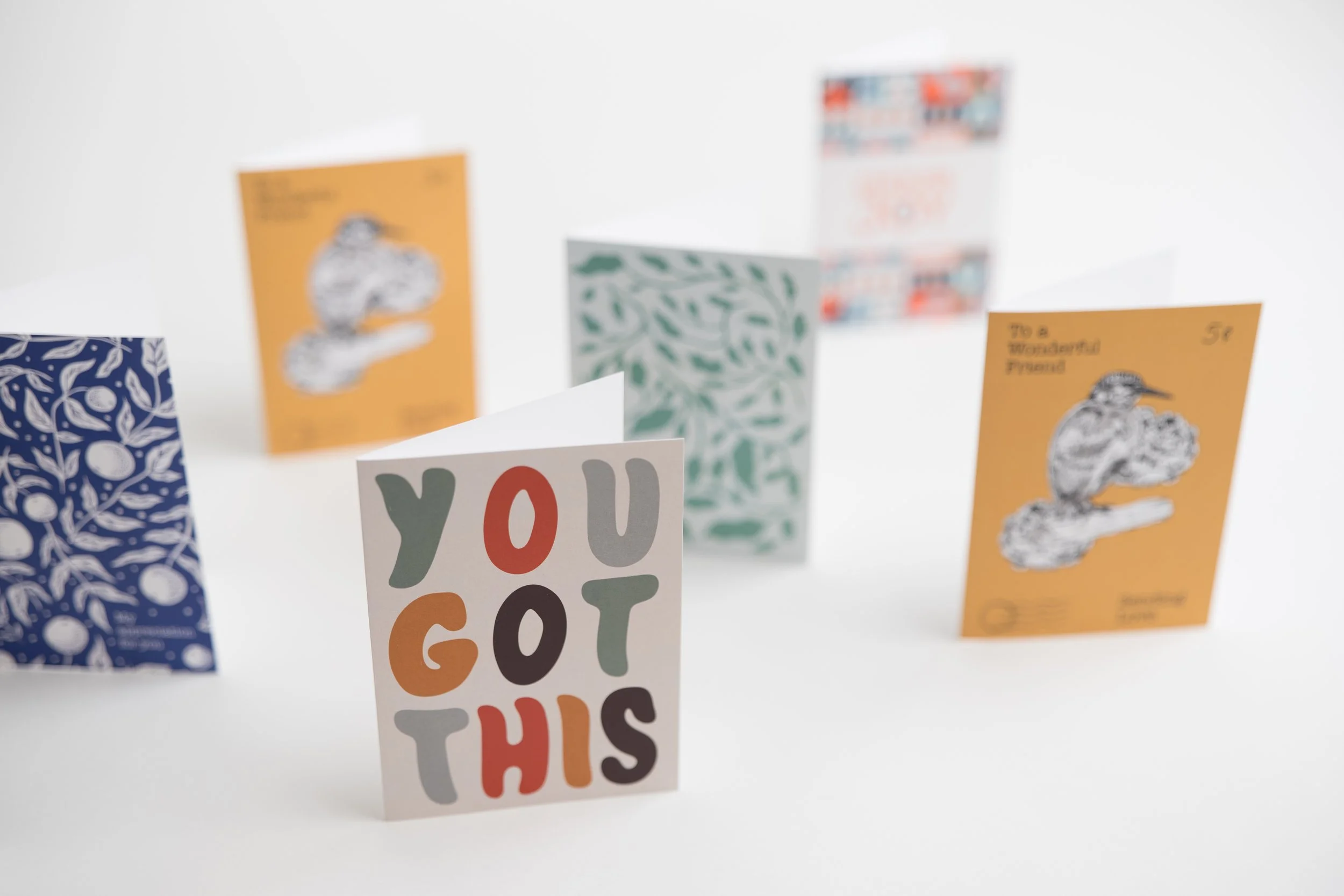 Multiple greeting cards arranged in a display, including one with colorful text saying "You Got This" and others with various designs and illustrations.