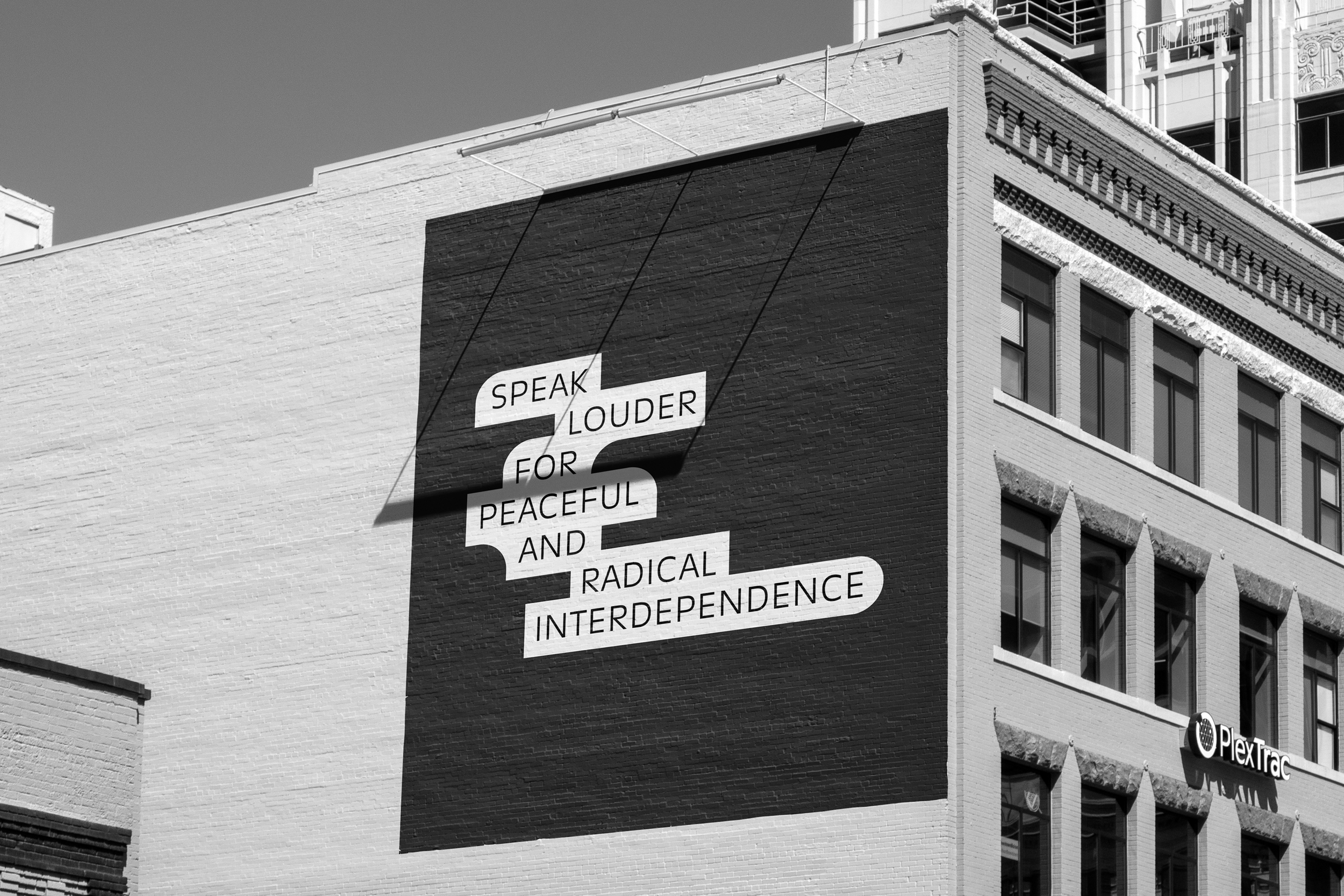 A large mural on the side of a building with the words "Speak louder for peaceful and radical interdependence" arranged in overlapping text on a black background in a city.