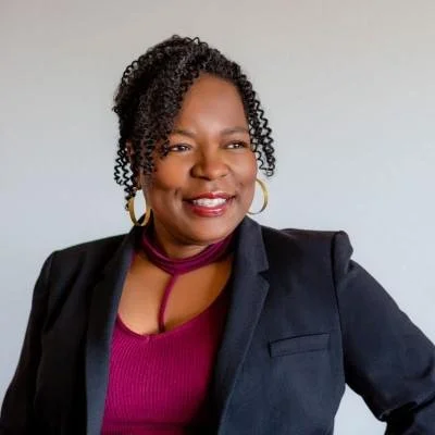 A Black woman with curly hair wearing a navy blazer, a purple top, gold hoop earrings,  smiling
