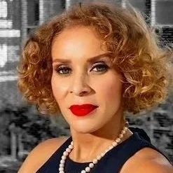 A Black woman with curly red hair wearing red lipstick, a pearl necklace, and a dark top, standing outdoors in front of an urban background.