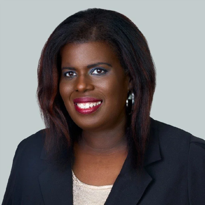 A Black woman with dark hair, red lipstick, and earrings, wearing a black blazer, smiling at the camera.