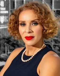 A Black woman with curly blonde hair wearing a navy blue sleeveless dress, pearl necklace, red lipstick, with an urban cityscape background.