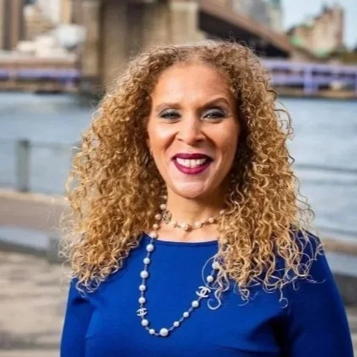 A Black woman with curly red hair wearing red lipstick, a pearl necklace, and a dark top, standing outdoors in front of an urban background.