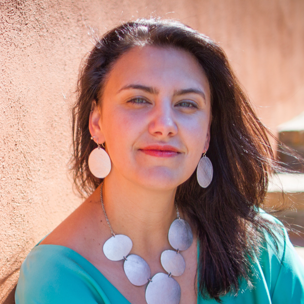 A white woman with dark hair wearing a turquoise top and large silver jewelry, posing outdoors against a peach-colored wall.
