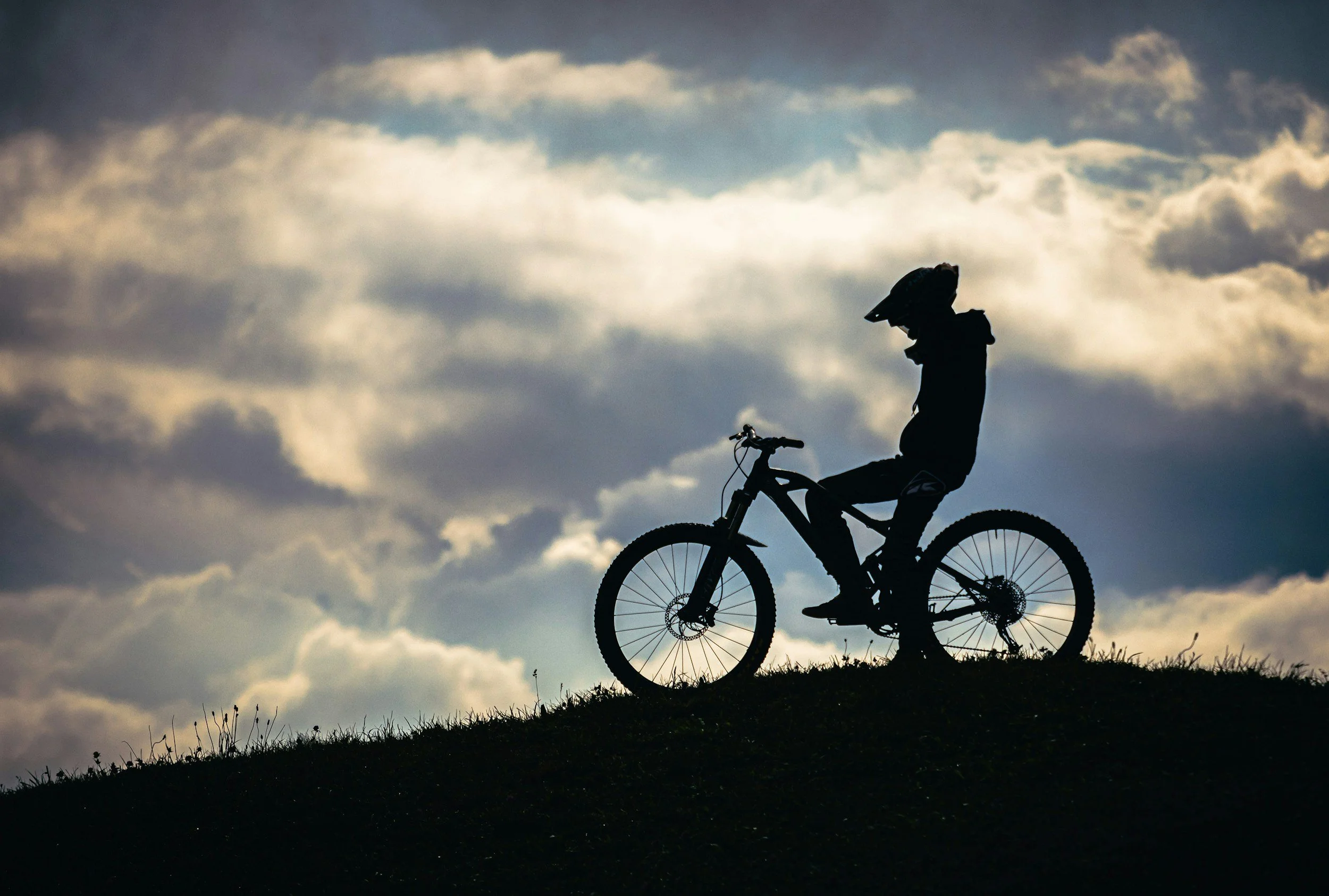 Silhouette of a person with a helmet riding a mountain bike on a hilltop against a cloudy sky during sunset.