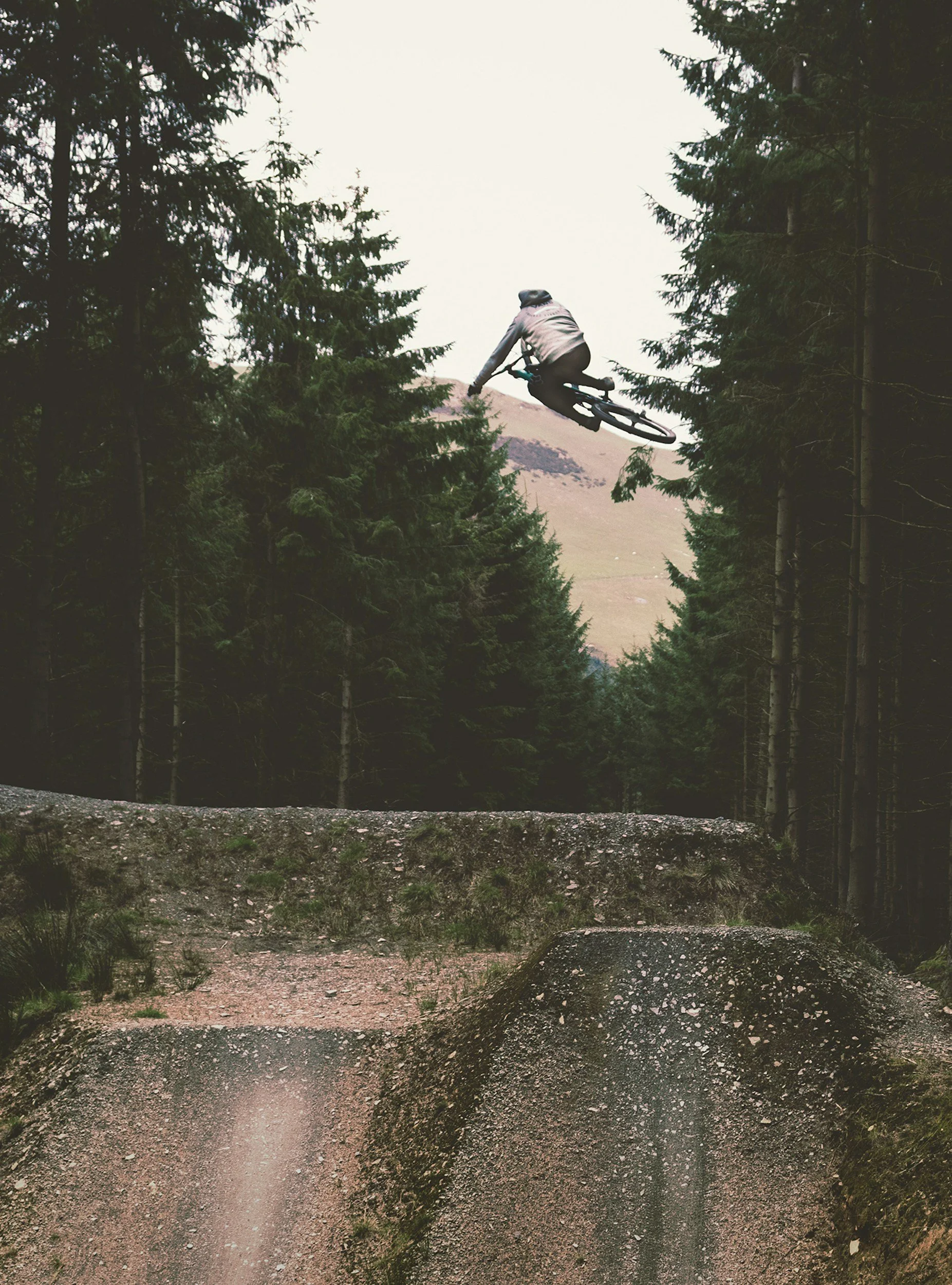 A person riding a mountain bike jumping off a dirt ramp in a forested area with tall trees and mountains in the background.