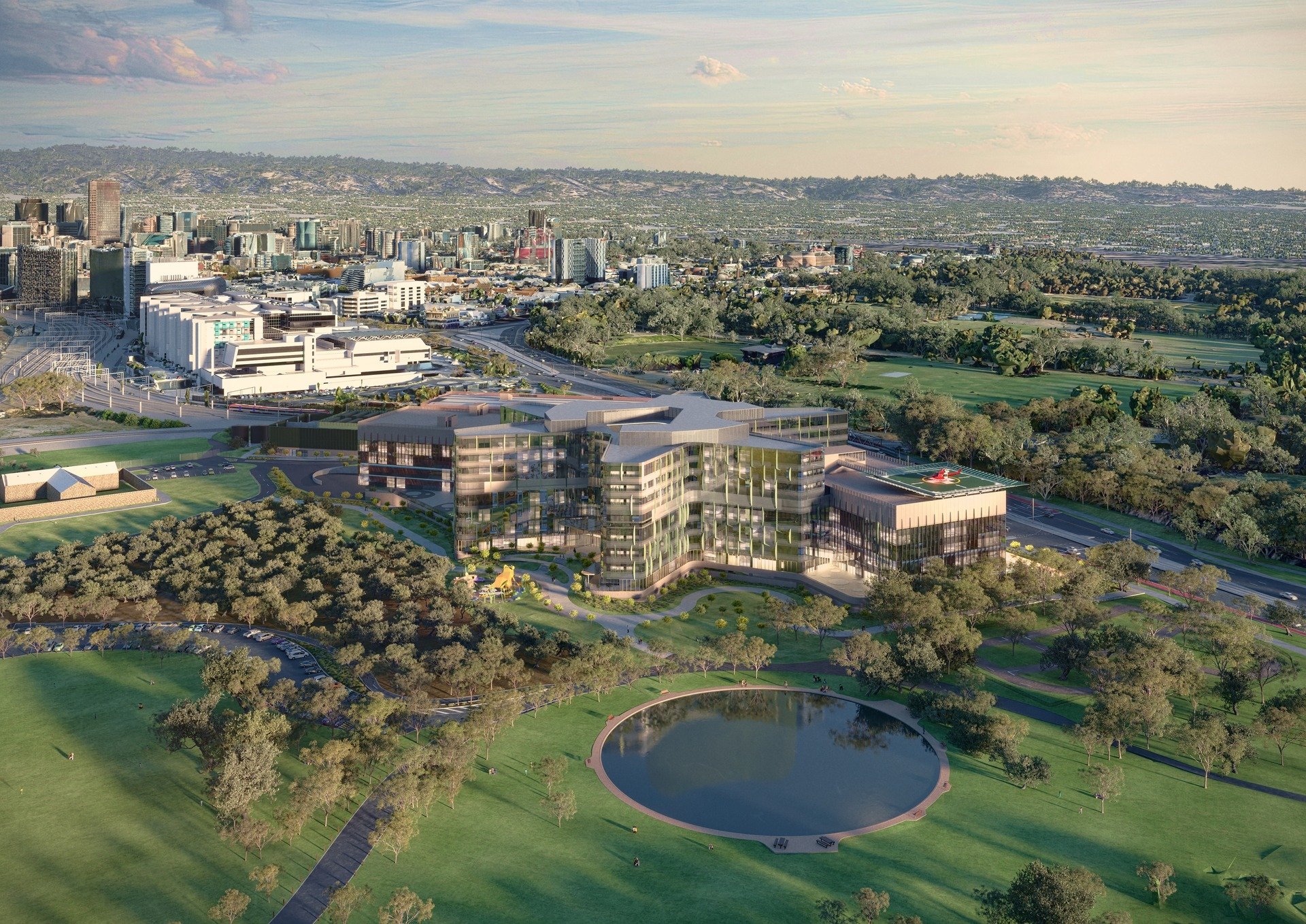 Aerial view of a modern building with green rooftop gardens, surrounded by trees, a pond, and open grassy areas with walking paths, with a city skyline in the background.