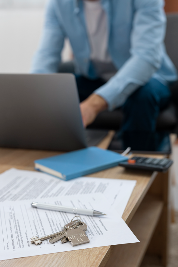 Close-up of a wooden desk with documents, a pen, keys, and a USB flash drive, with a person in a blue shirt sitting in the background using a laptop.
