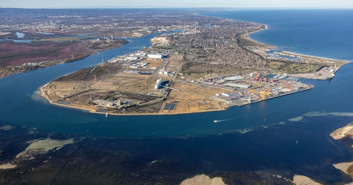 Aerial view of a coastal industrial area with shipping containers, warehouses, and a bridge over a river, with a city and ocean in the background.
