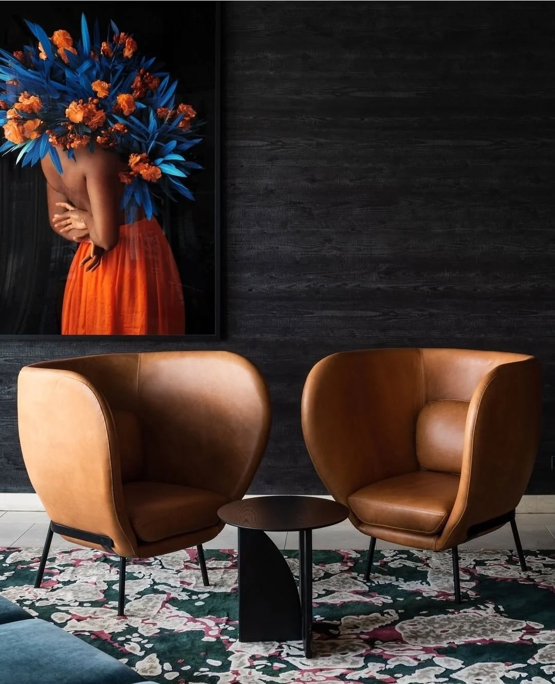 Sala de estar con dos sillones de cuero marrón, una mesa negra en medio y una alfombra con diseño floral. En la pared hay un cuadro con una imagen de una persona con un atuendo color naranja y un tocado de flores azules y naranjas.