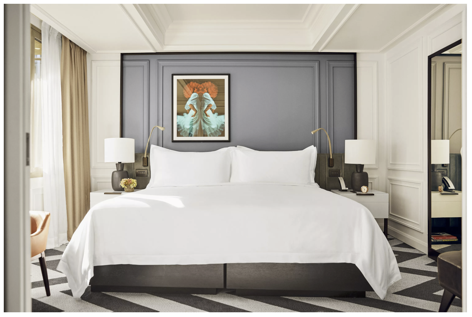 Habitación de hotel con cama grande, sábanas blancas, dos lámparas, cuadros y una pared azul grís con cortinas beige y blancas, espejo y una silla naranja a la izquierda.