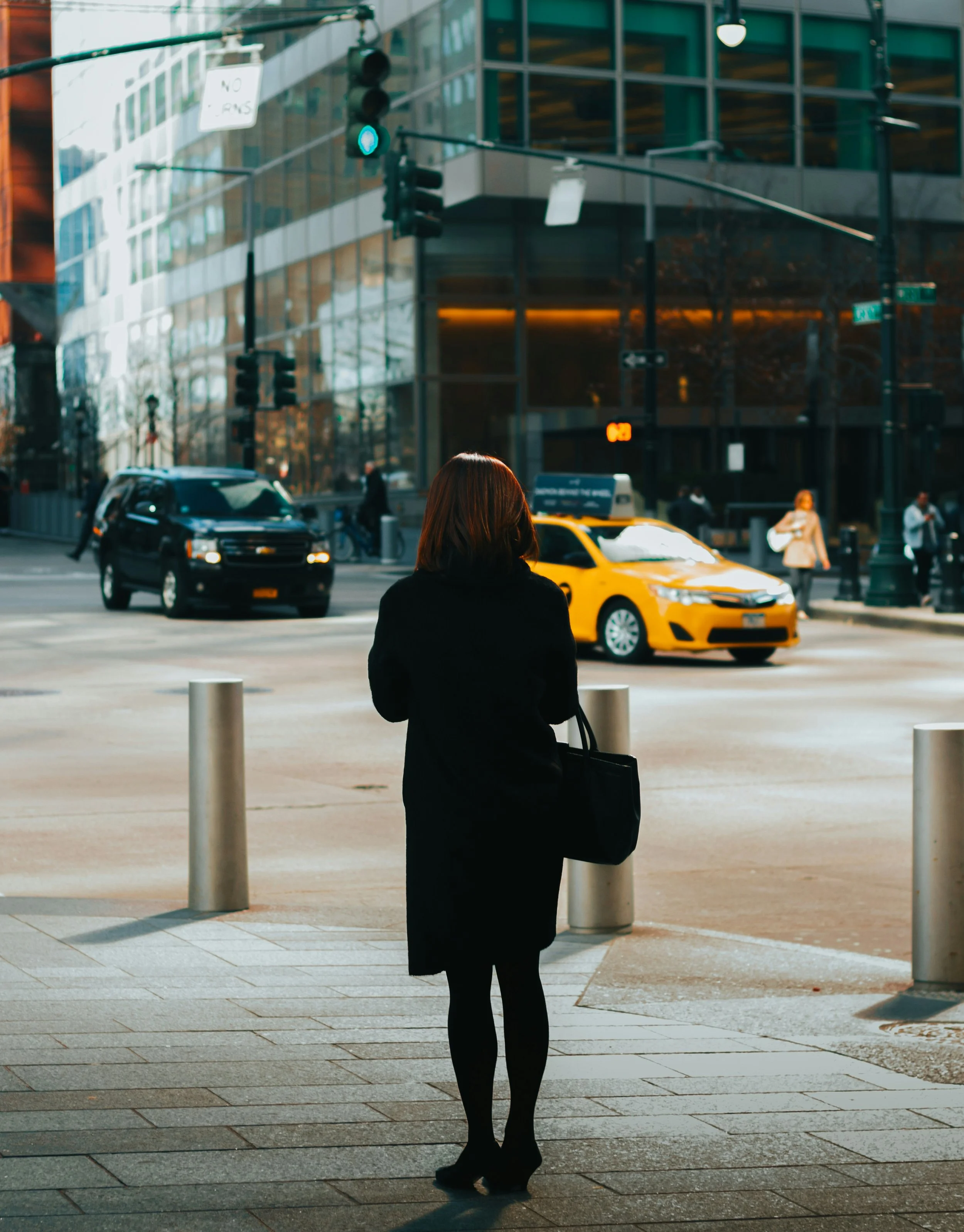 A woman in black clothing and heels, carrying a black bag, stands at a city intersection waiting to cross as cars, including a yellow taxi and a black SUV, pass by. Modern glass buildings reflect on the street.