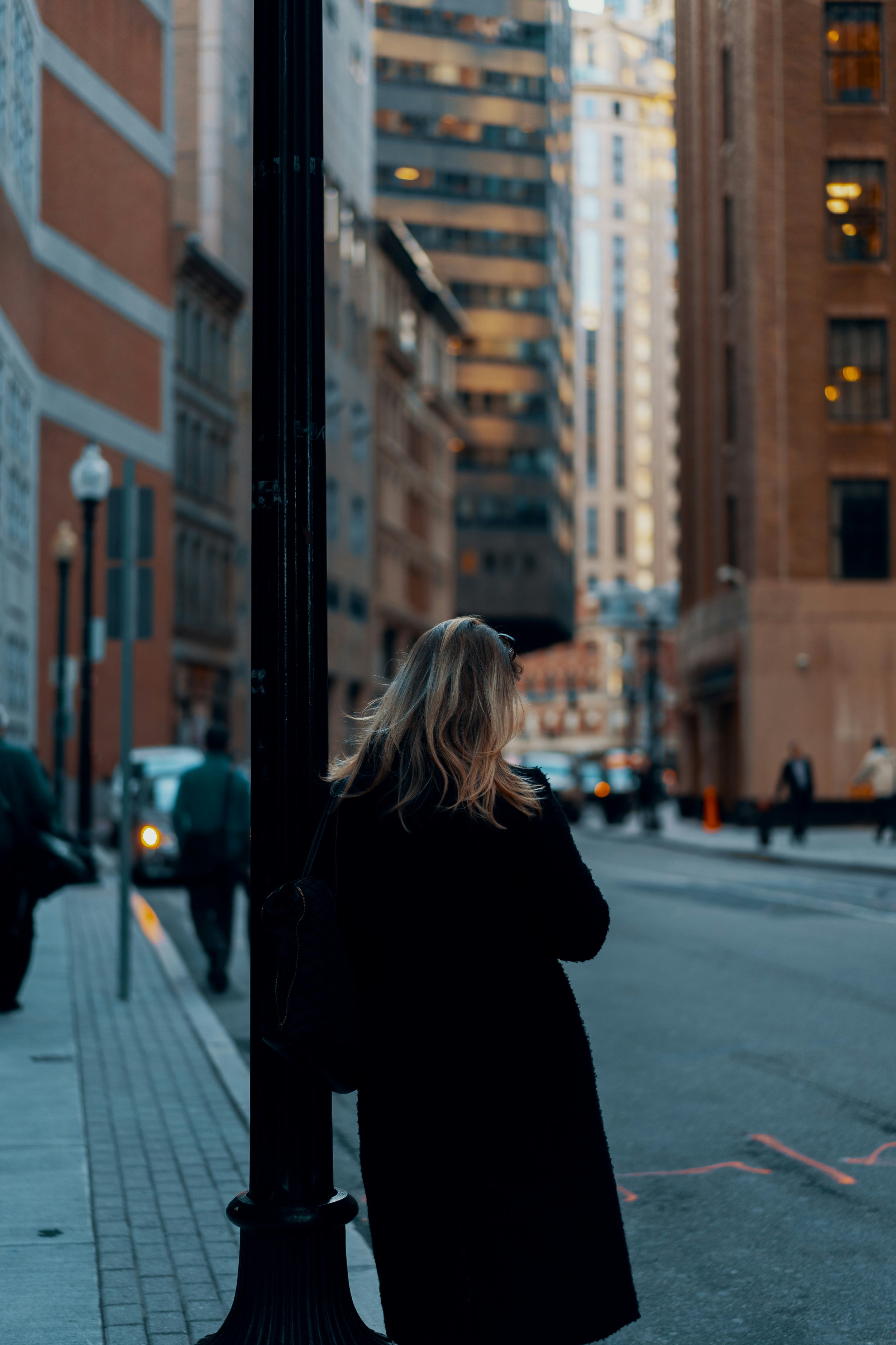 A woman with blonde hair wearing a black coat stands on a city sidewalk, leaning against a black lamppost, with tall buildings and a street in the background.