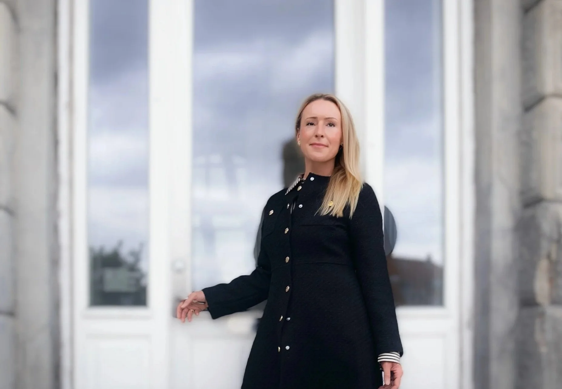 A woman with blonde hair wearing a black dress with gold buttons standing in front of a large window with a cloudy sky outside.