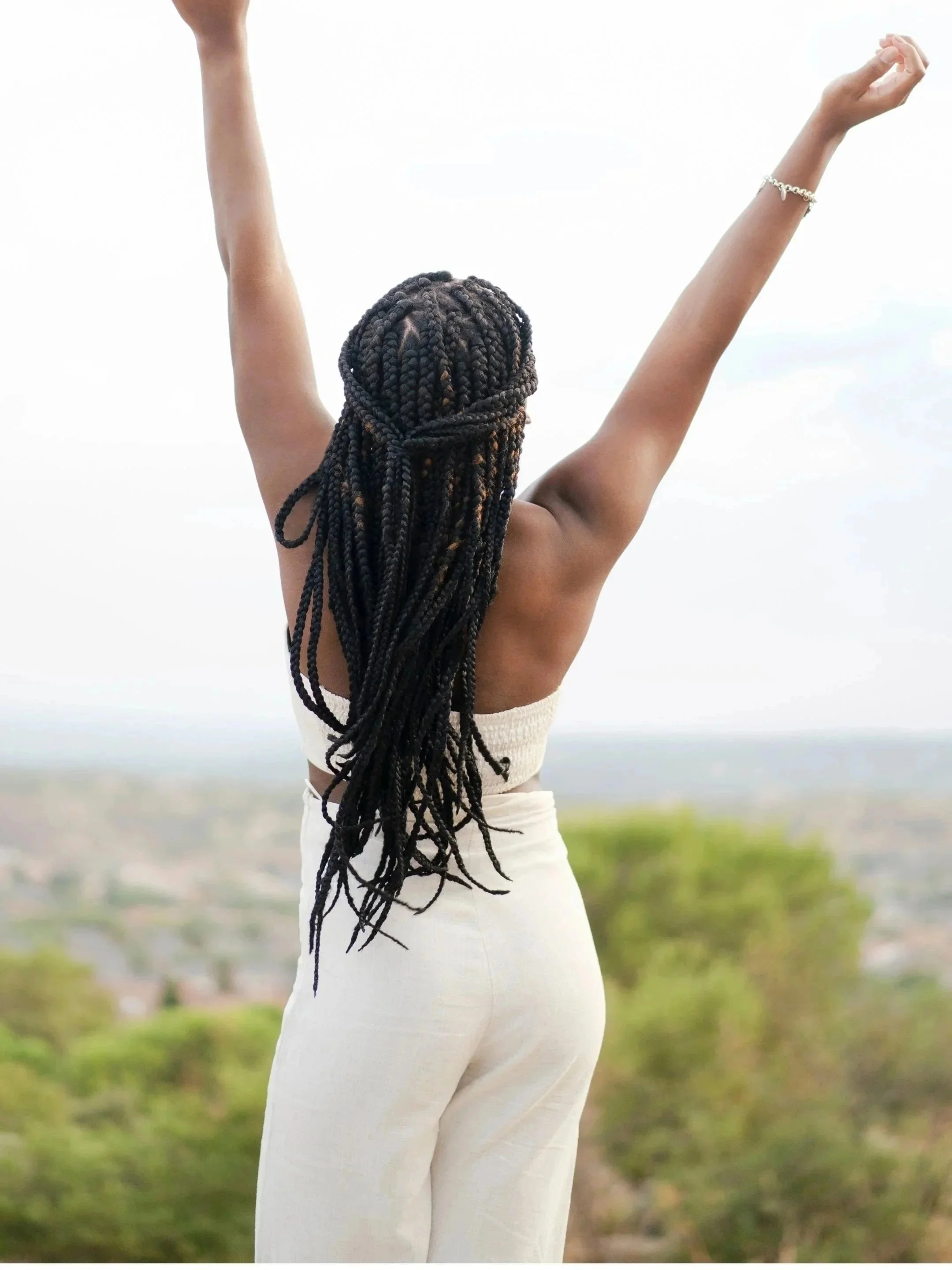 A woman with long braided hair standing outdoors with arms raised, facing away from the camera, in a light-colored outfit.