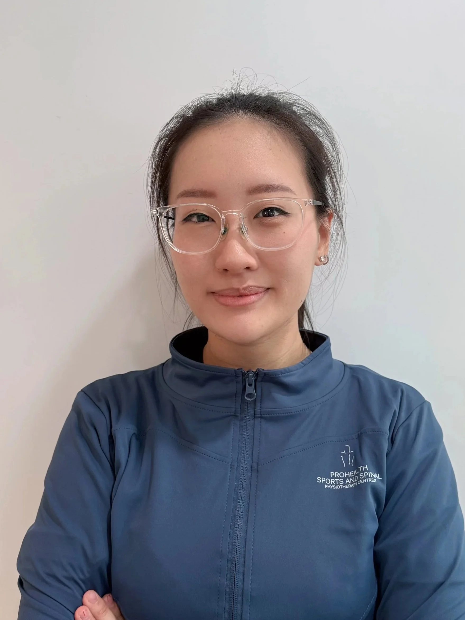 A woman with glasses and straight dark hair wearing a blue athletic jacket standing against a plain white background.